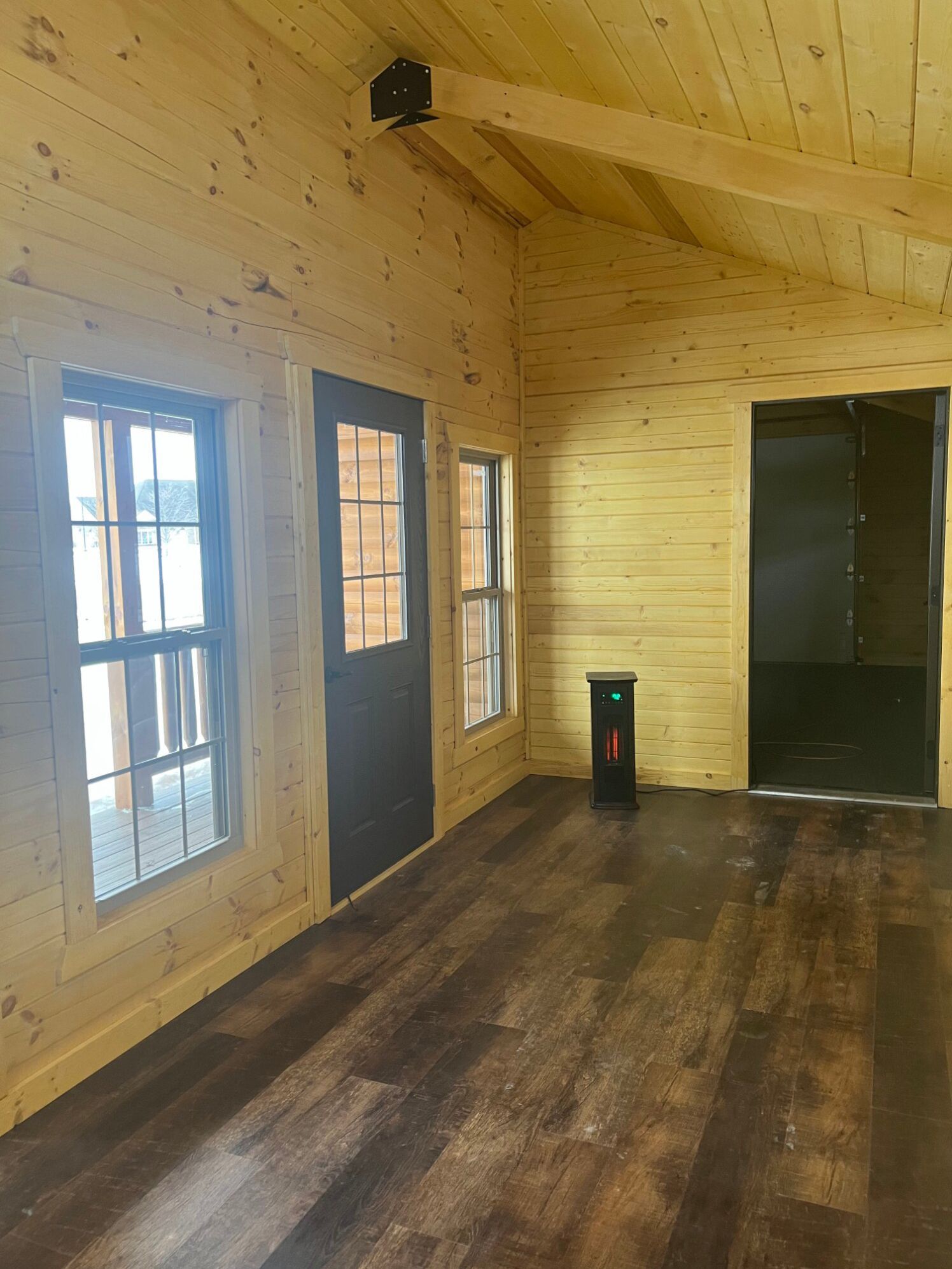 Interior view of a rustic cabin with wood-paneled walls, dark wood flooring, a door, two windows, and an open doorway.
