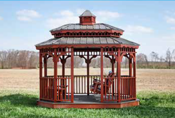 Red wooden gazebo in a grassy field with a person seated inside, under a blue sky.