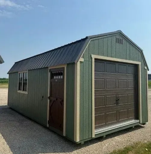 Green shed with dark brown door and garage door. Tan trim, black roof, gravel ground.