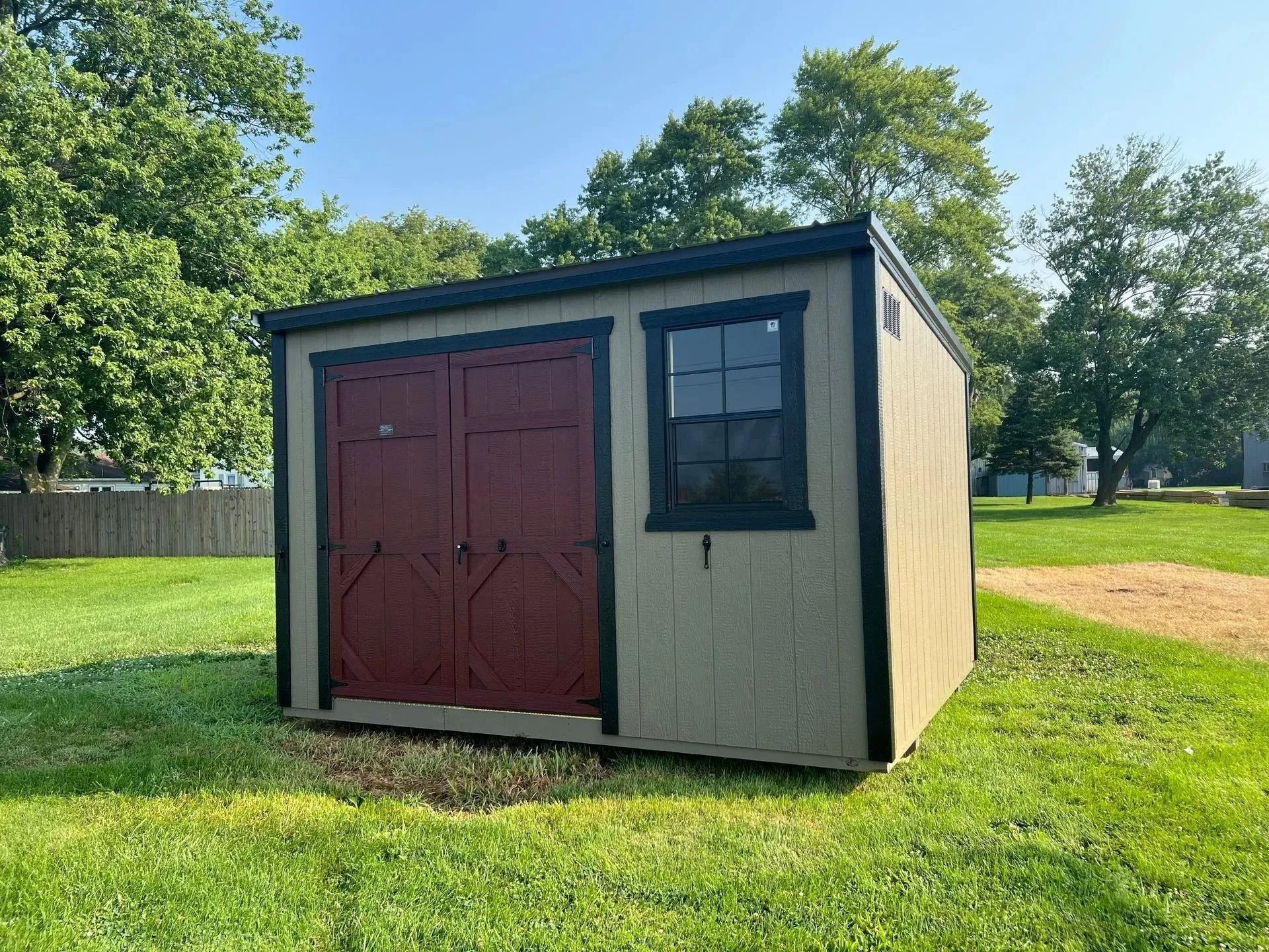 Small tan shed with red double doors and a black window, standing on a grassy lawn.