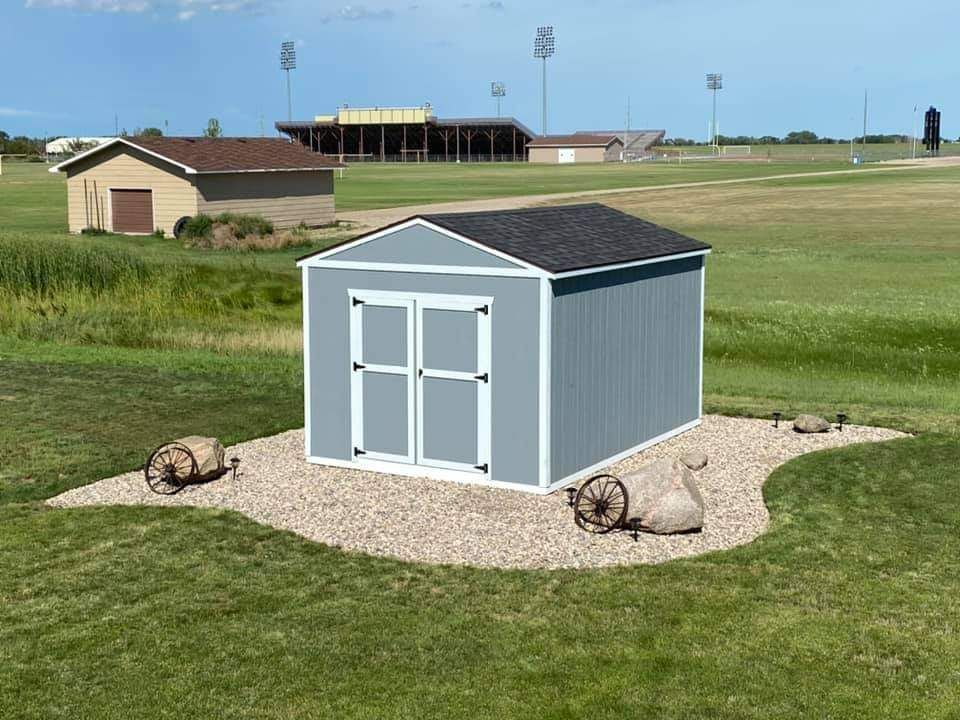 Blue shed with white trim, set on gravel, on a grassy field, with a ball field in the background.