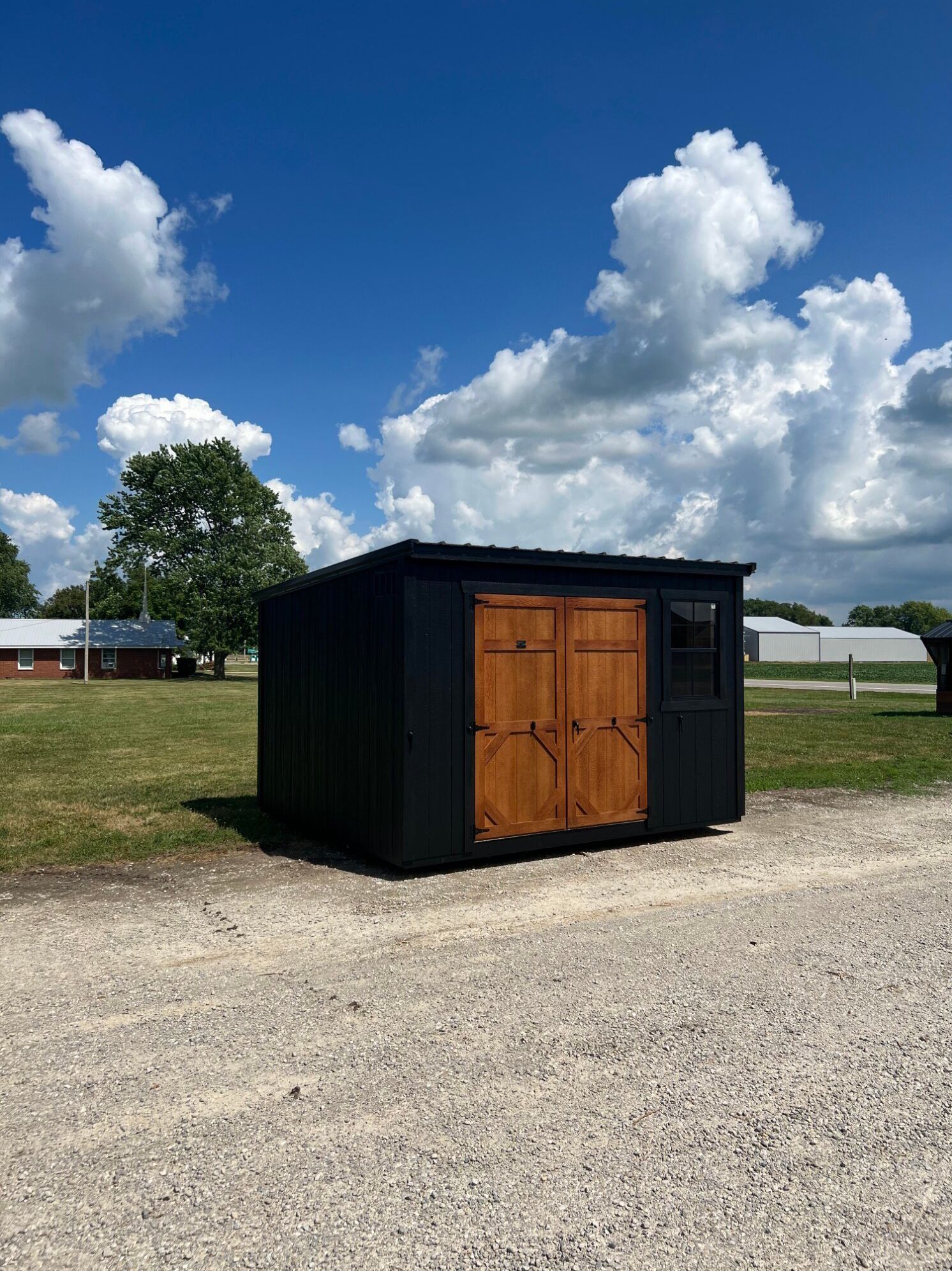 Small black storage shed with wooden double doors on a gravel lot under a blue sky with clouds