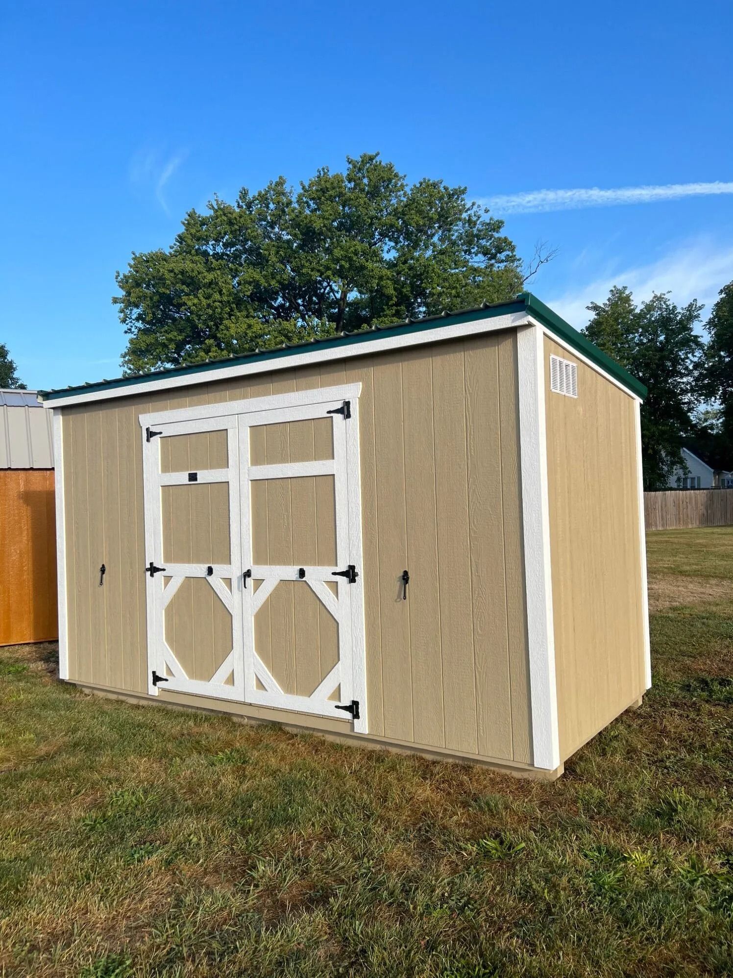 Small tan storage shed with white double doors on a grassy yard under a blue sky