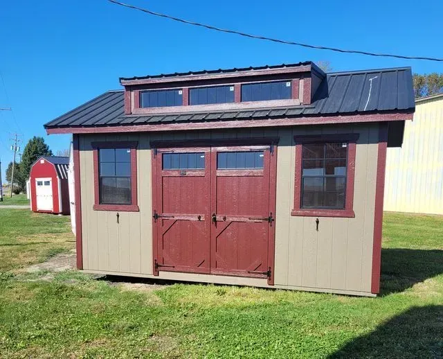 Tan shed with maroon trim and doors, black roof, and windows against a blue sky.