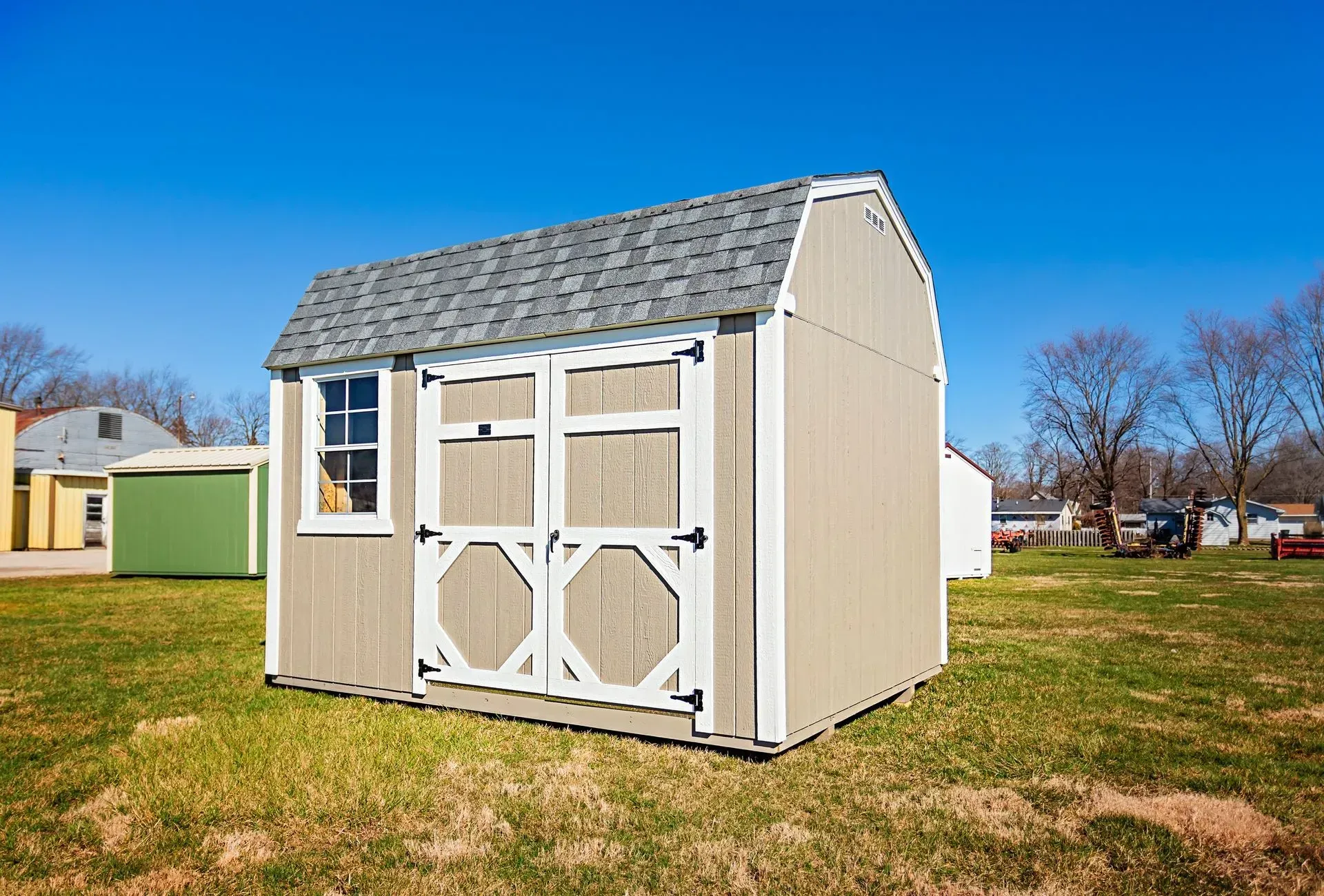Tan shed with gray roof and white trim in a grassy yard, under a blue sky.