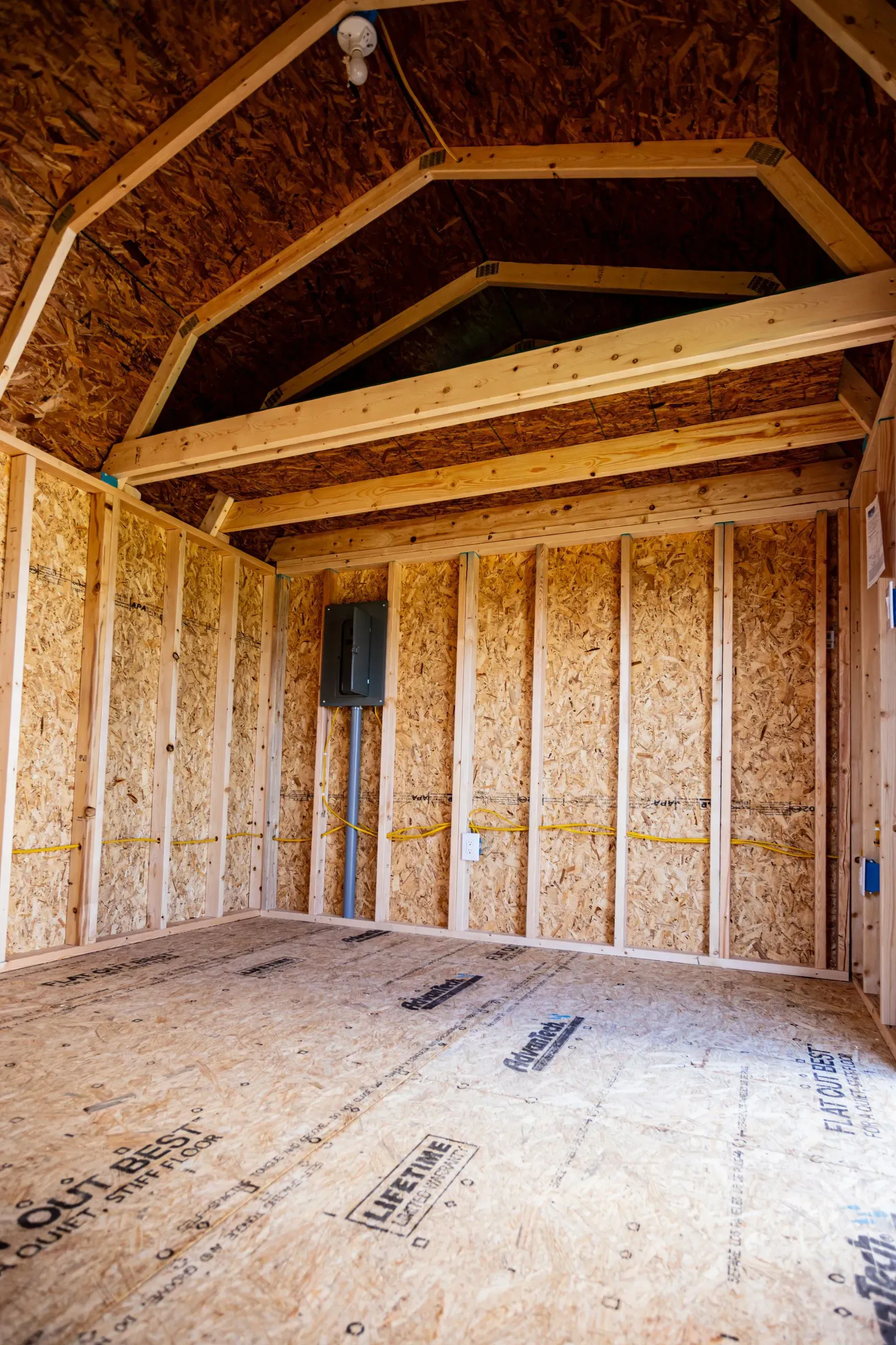 Interior view of a new, unfinished shed with exposed wood framing, walls, and ceiling, with a light fixture and electrical box.
