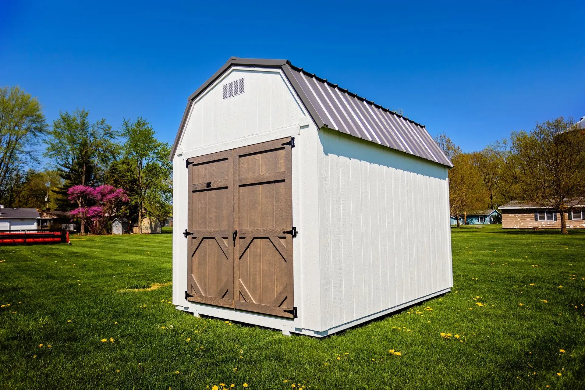 White shed with brown double doors, gray roof, on green grass under a blue sky.