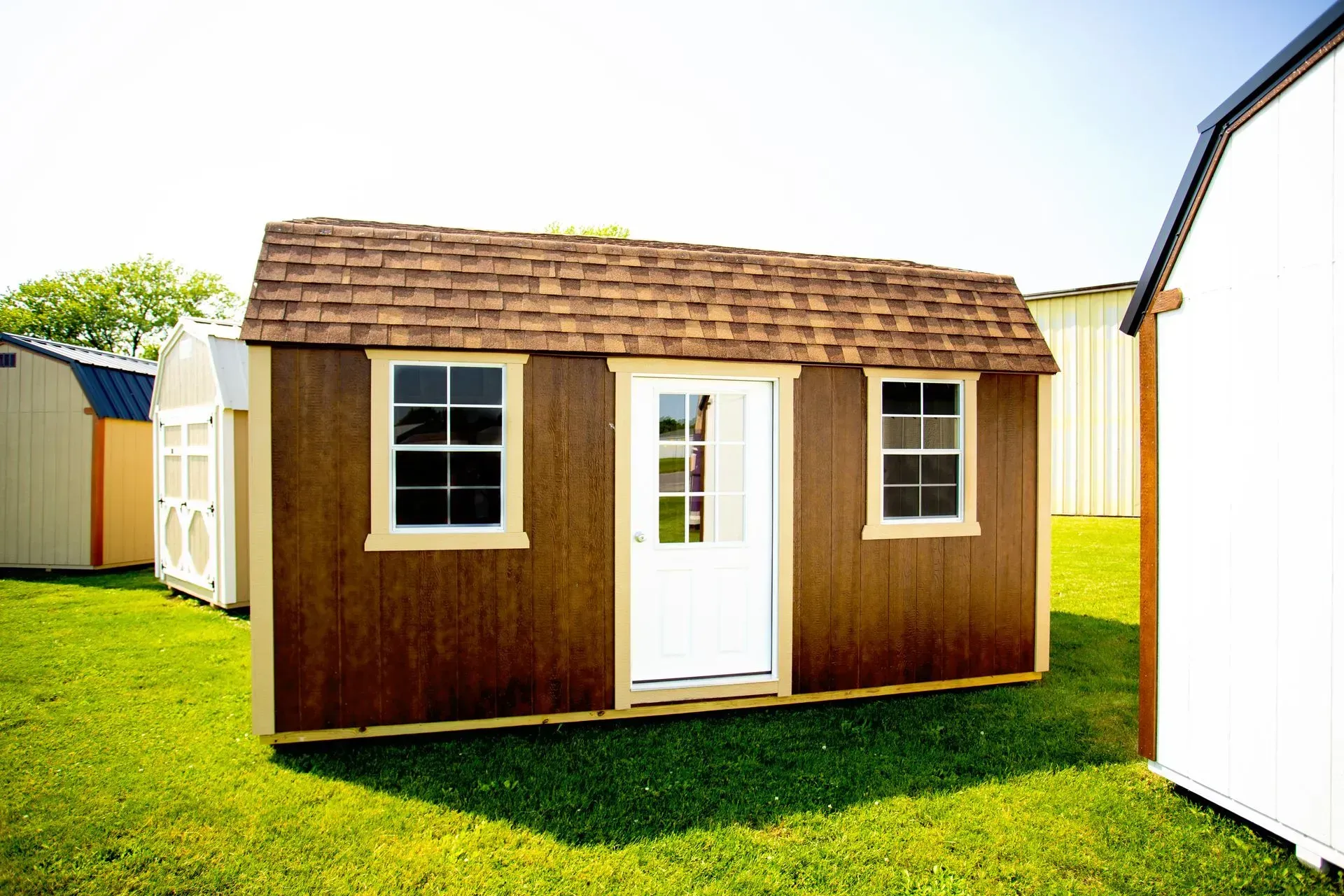 Brown shed with a white door and two windows on green grass.