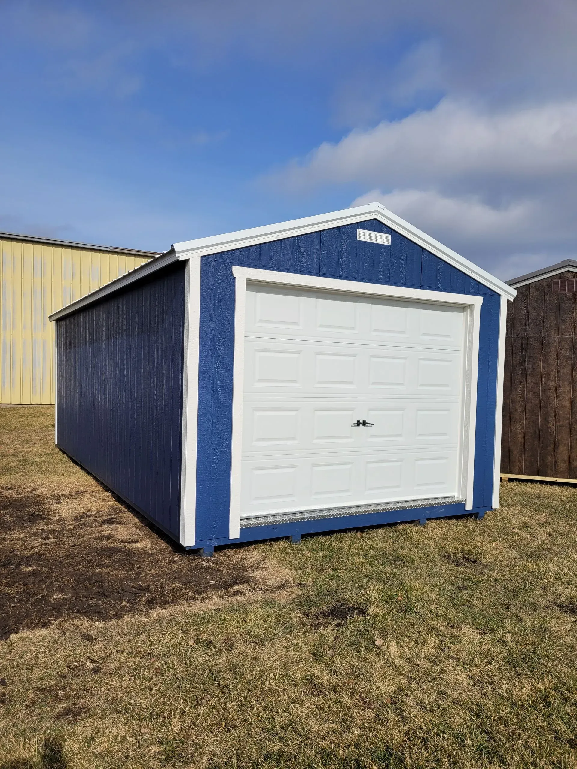 Blue shed with white trim and a white garage door, set on brown grass against a blue sky.