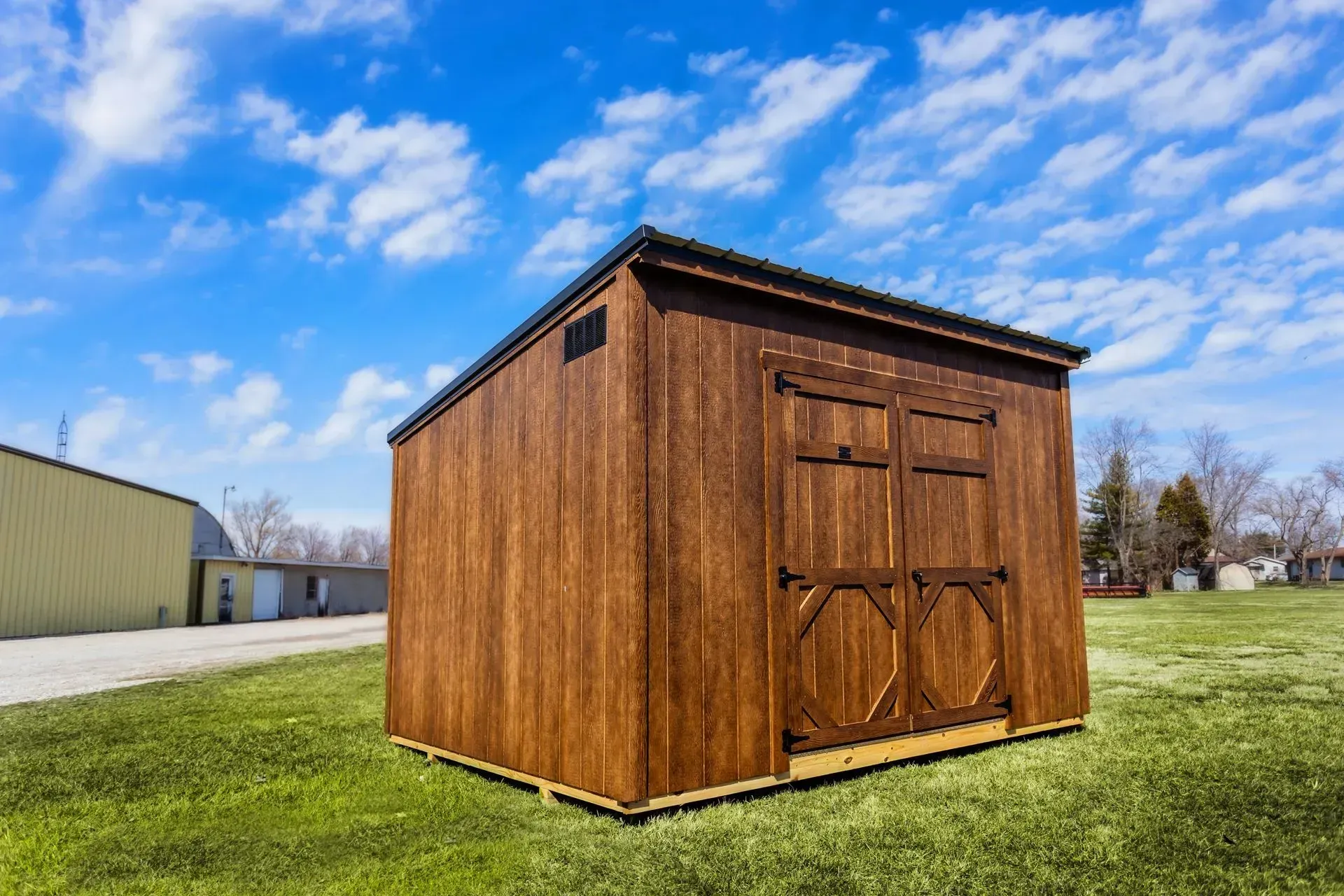 Brown wooden shed on a grassy field under a blue sky with clouds.