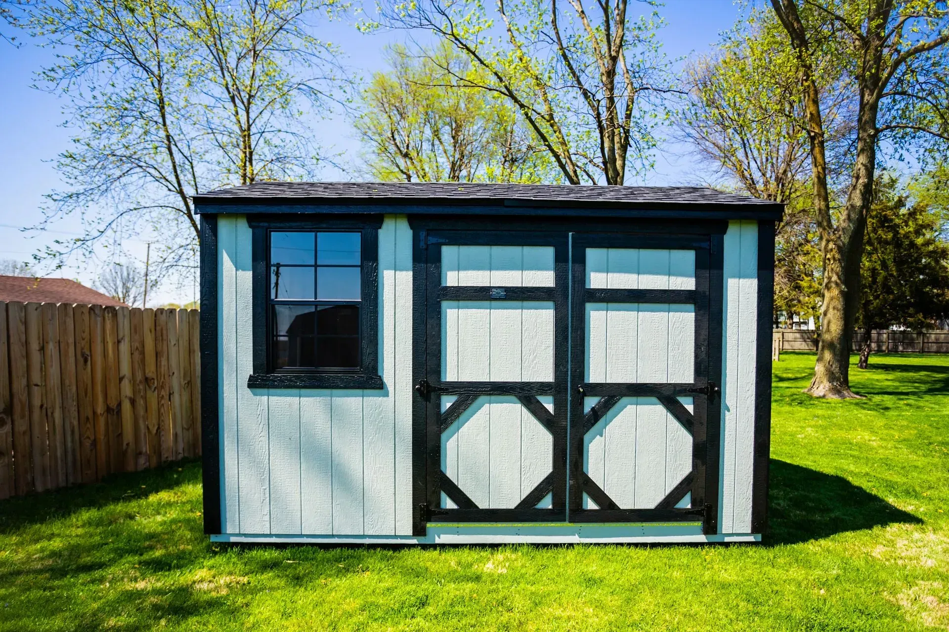 A small, light blue shed with black trim and a window sits on a green lawn with a wooden fence and trees in the background.