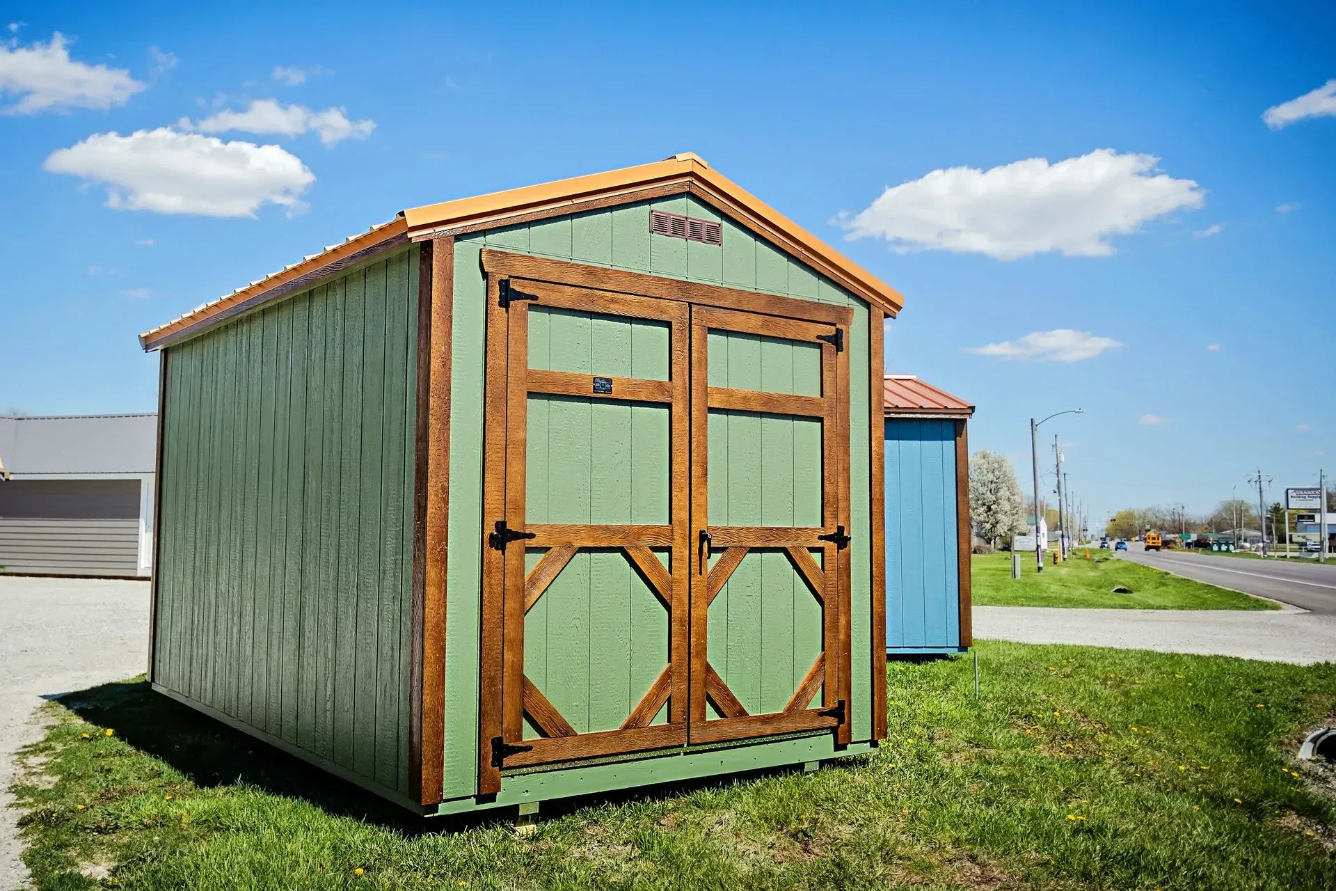 Green wooden shed with brown trim, doors, and roof against a blue sky.
