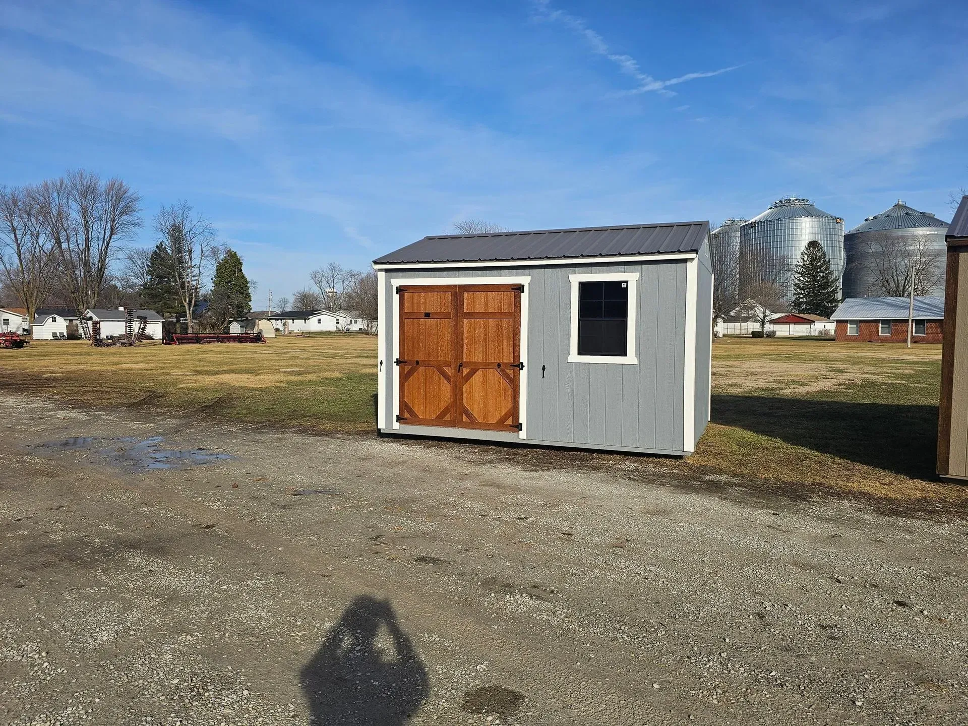 Gray shed with brown doors and a window, gravel lot, silos and houses in background.