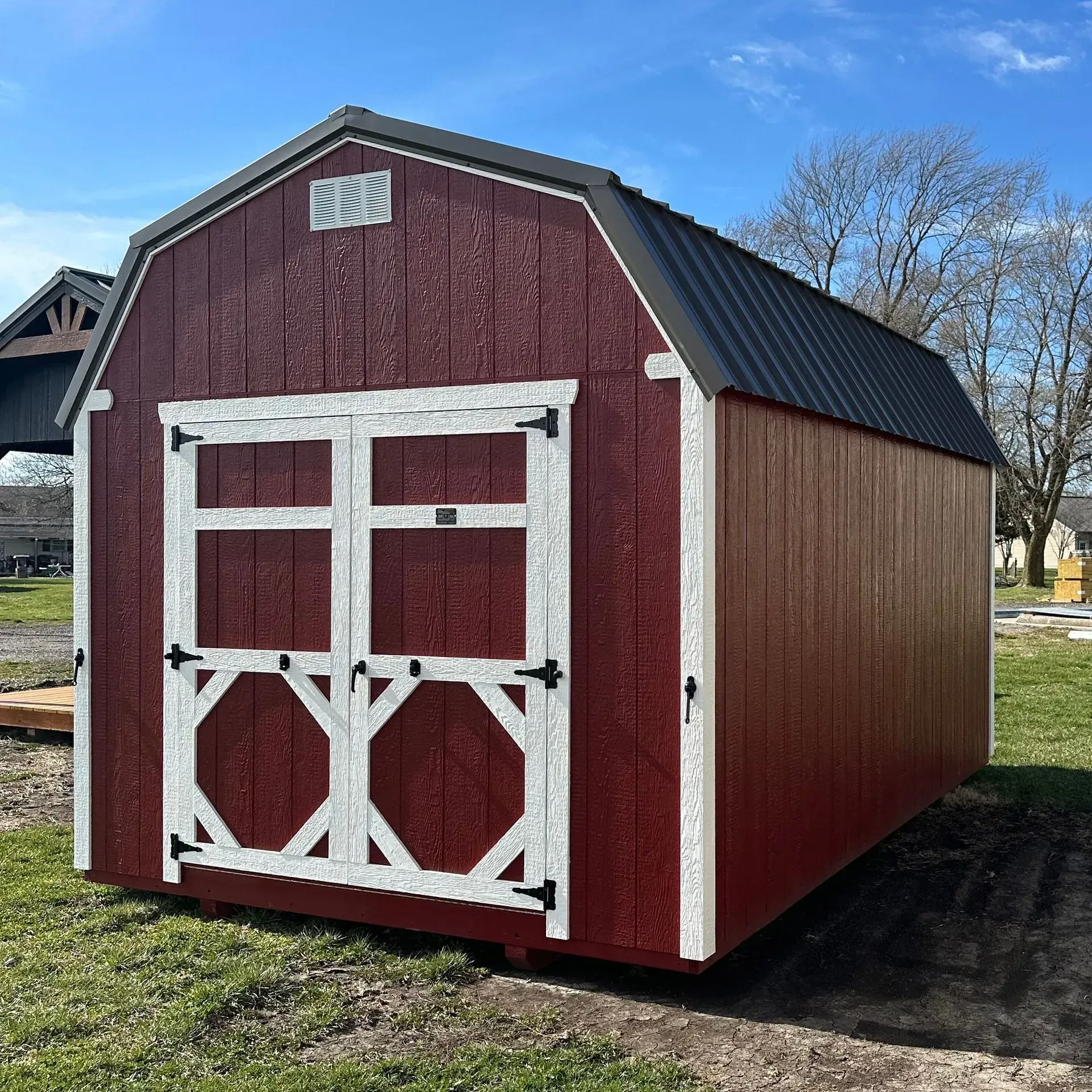 Red barn-style shed with white trim and double doors; black metal roof on green grass.