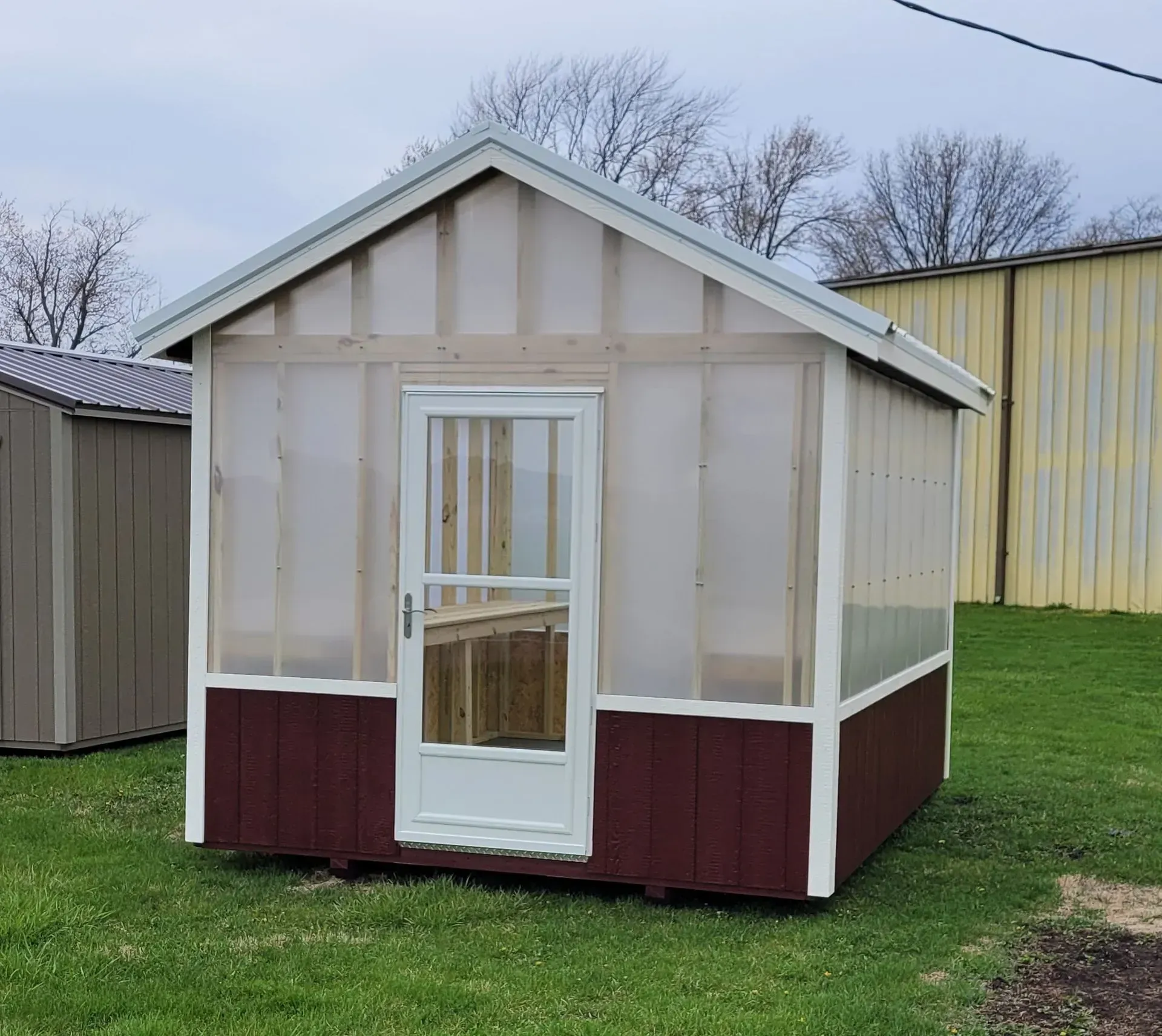 A small, enclosed greenhouse with a white door and translucent walls and a burgundy base sits on green grass.