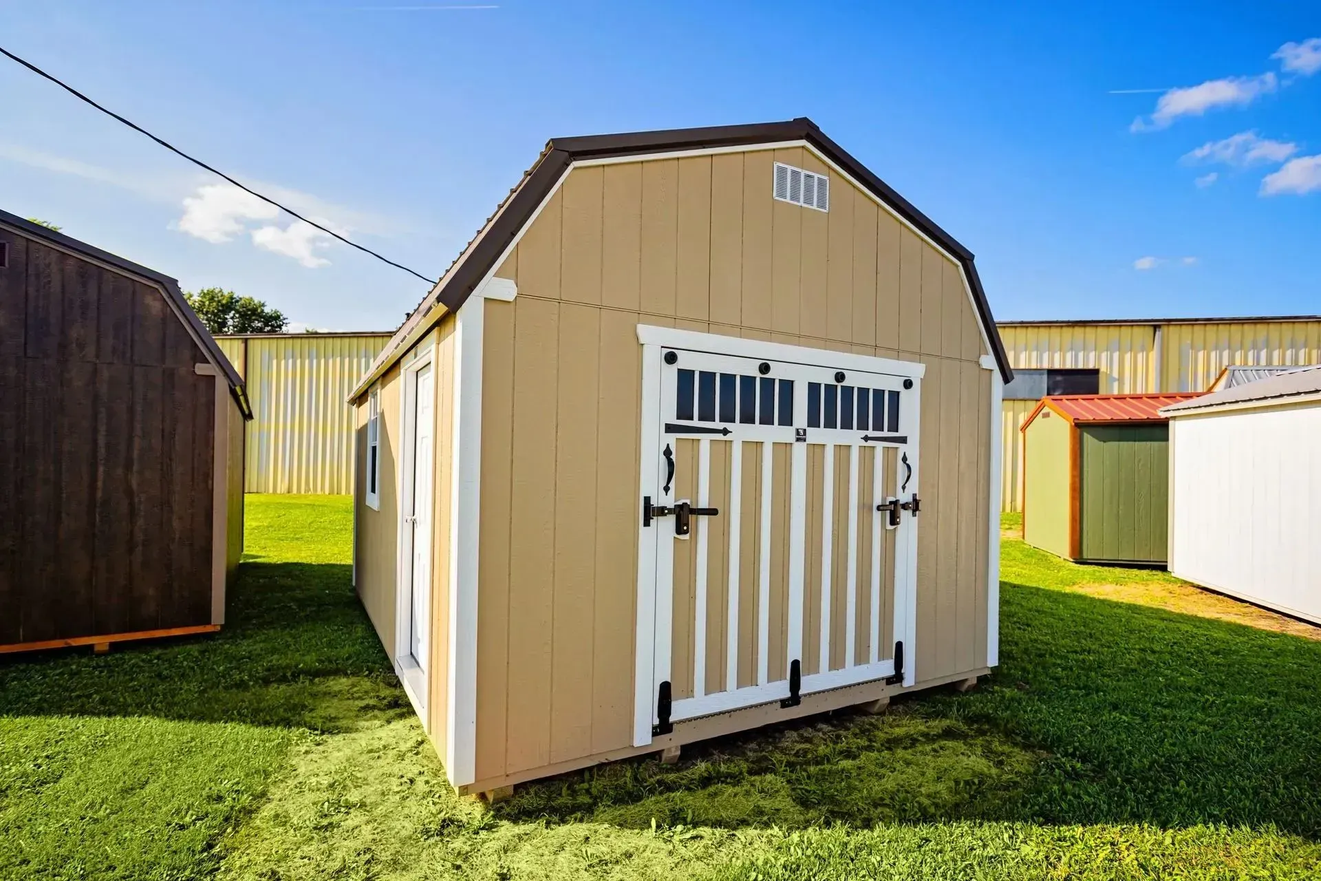 Tan shed with white trim and barn-style doors, set on grass against a blue sky.