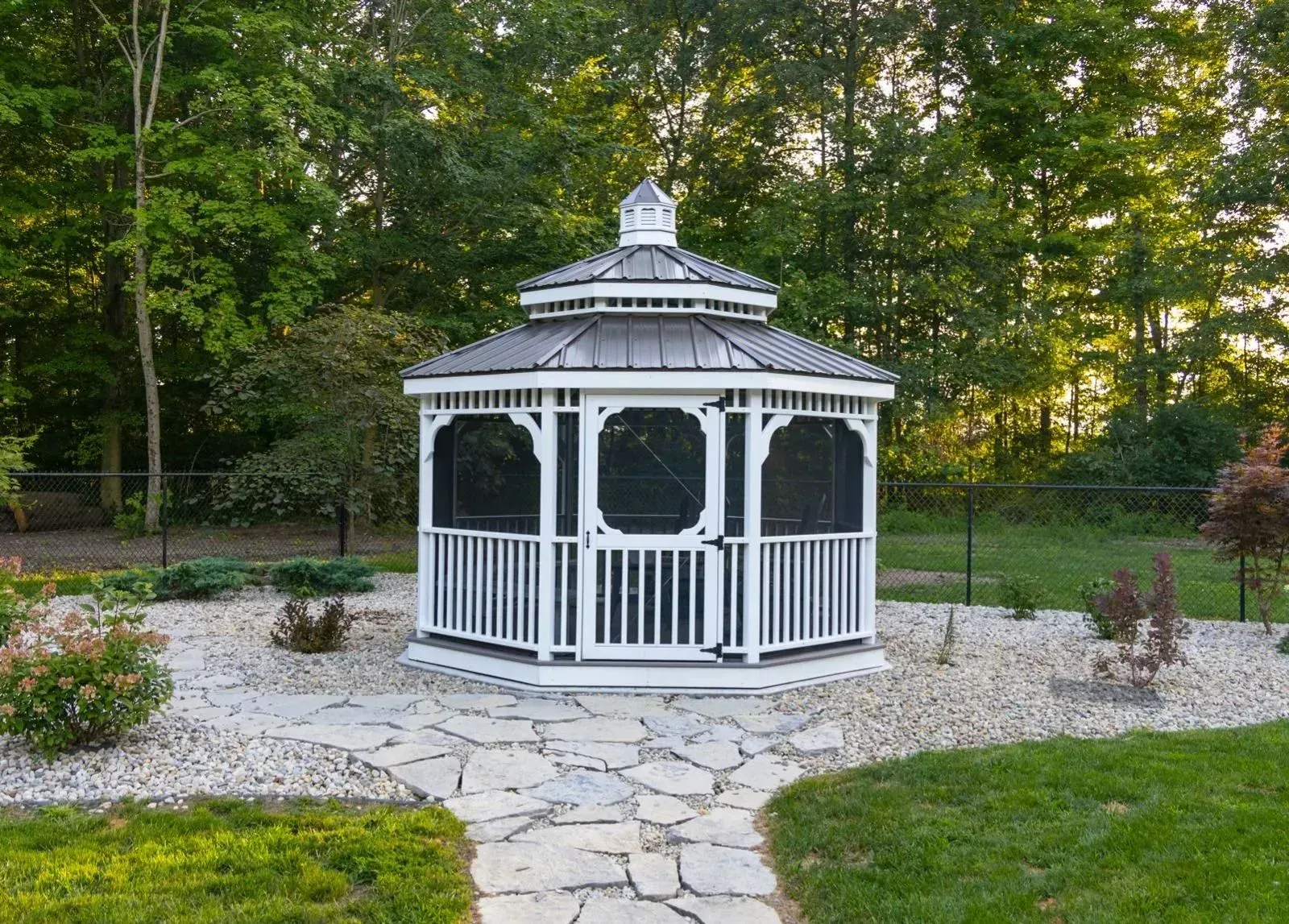 White gazebo with screen windows and decorative roof in a stone garden, surrounded by trees and grass.