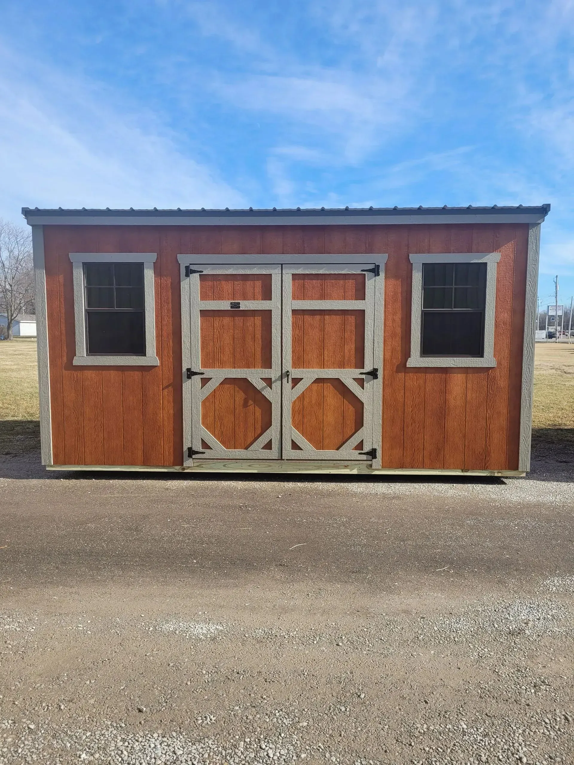 Brown shed with double doors, two windows, set on a gravel surface under a blue sky.