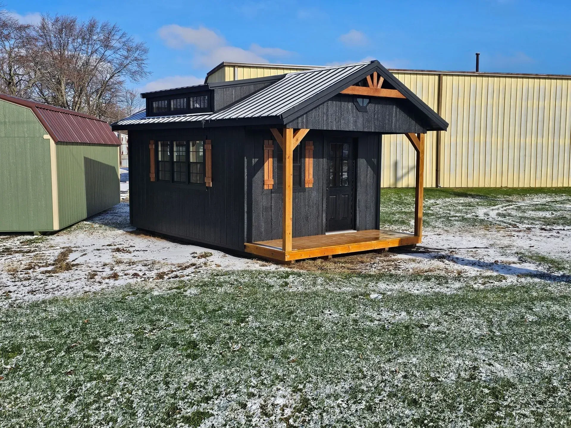Black cabin with wooden porch and shutters sits on snowy ground.