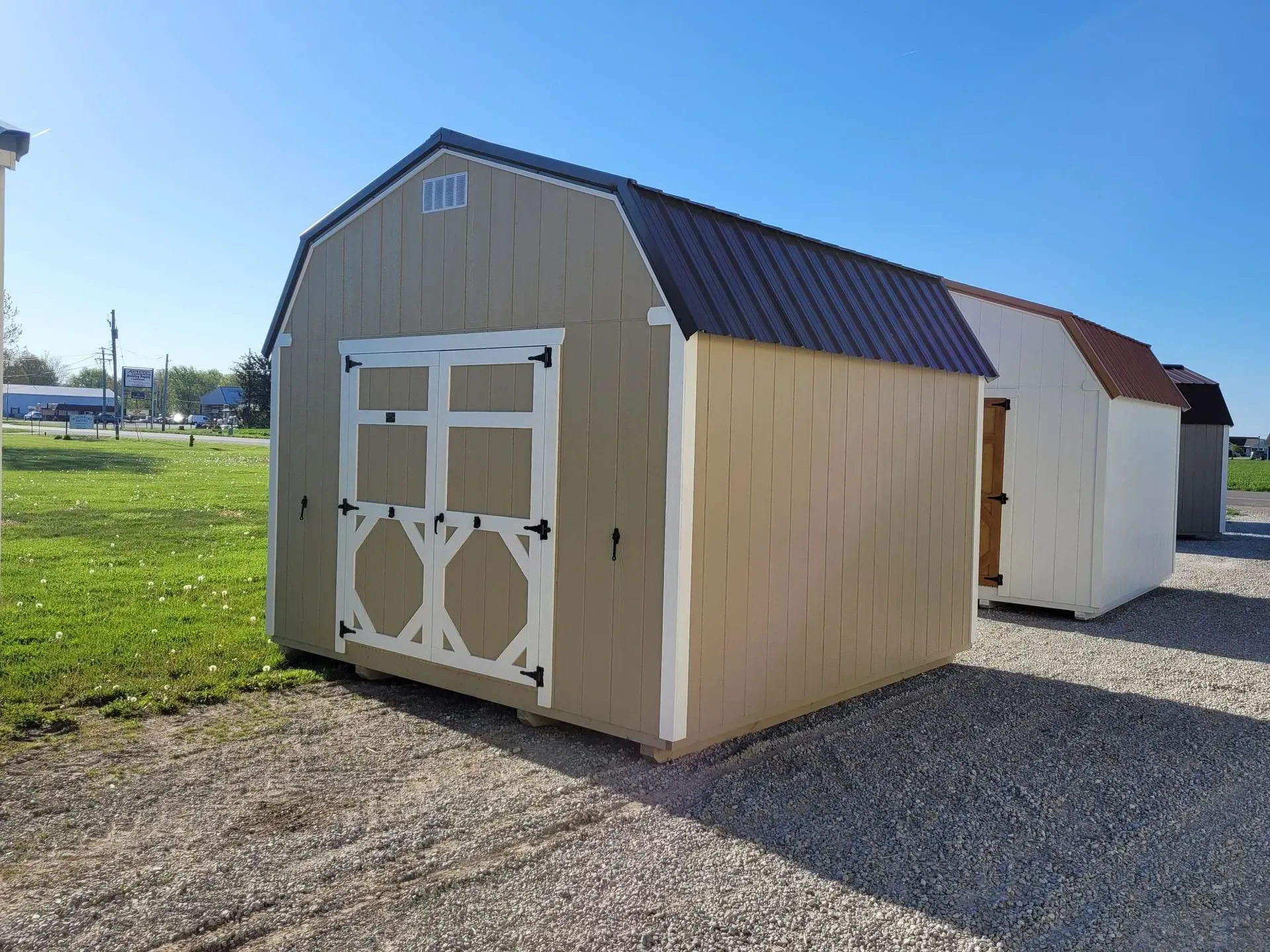Tan shed with dark roof, white trim, and a second shed visible in background, outdoors on gravel.