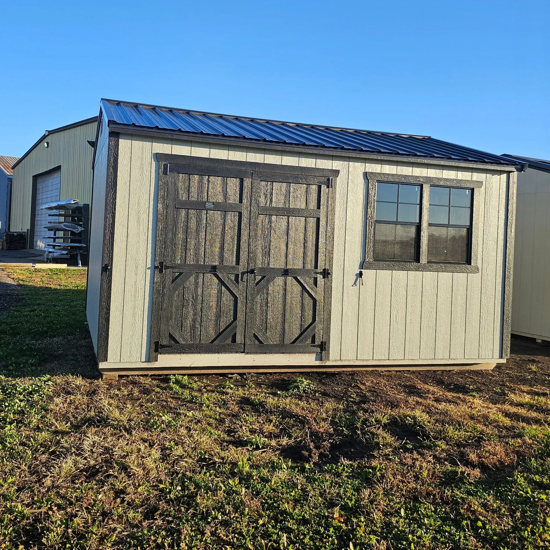 Tan shed with black doors and window; black roof on a grassy field.