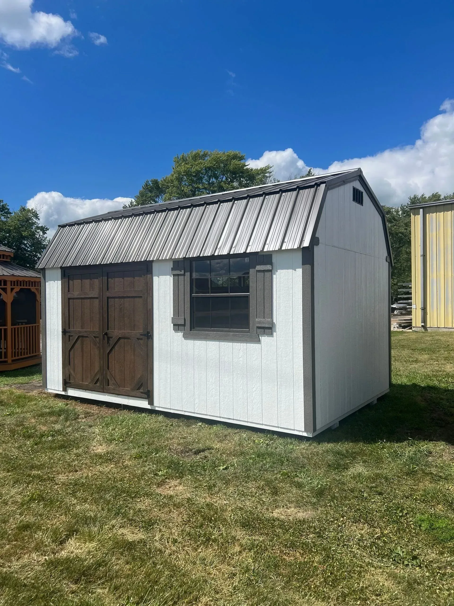 White shed with brown doors and trim, a small window, and a wood shake roof sits on grass under a blue sky.