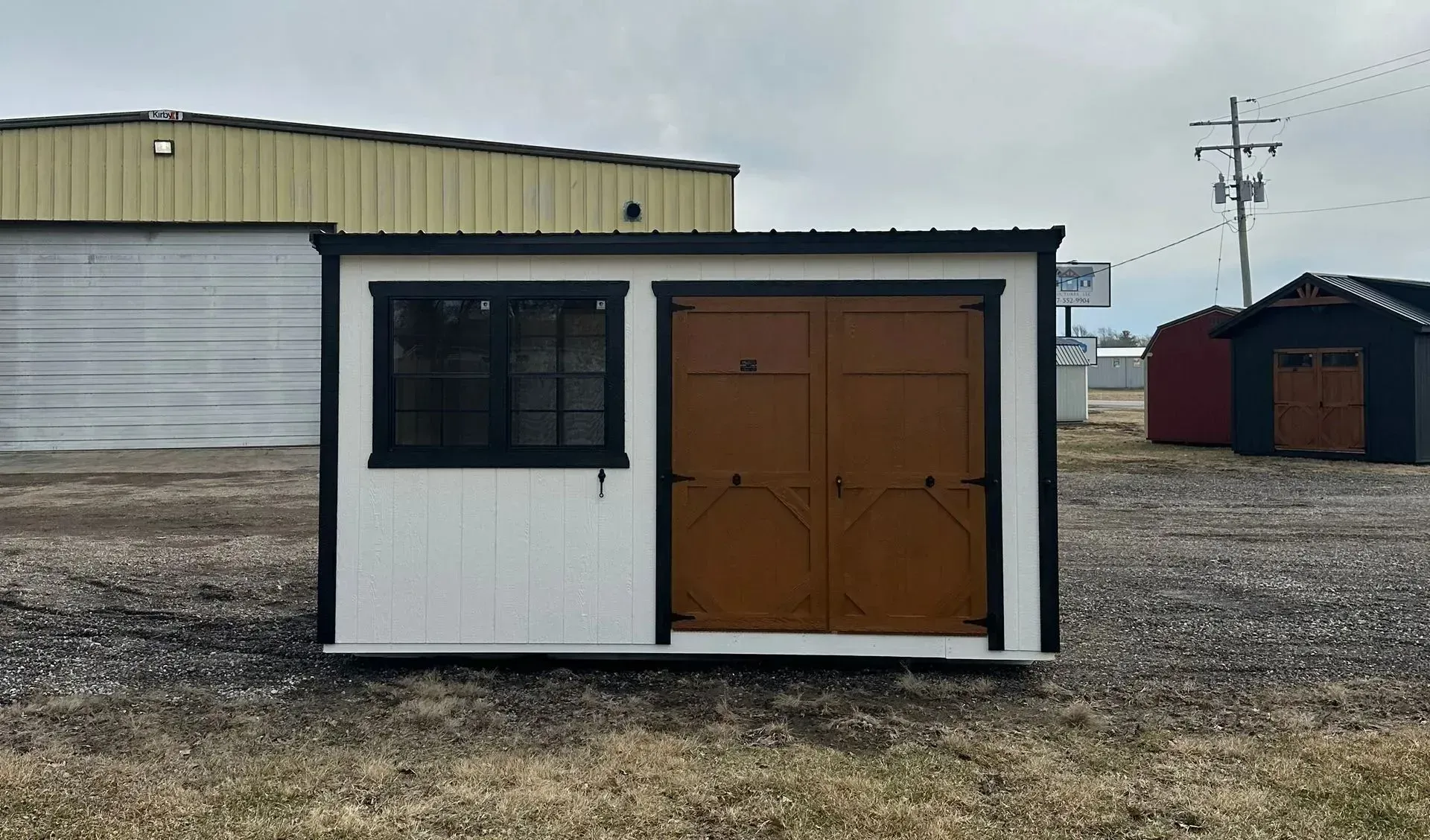 White shed with a brown door and black trim, window on the left, set on gravel, cloudy day.