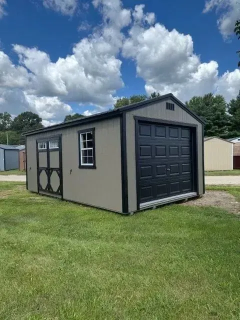 Tan shed with black garage door, window, and trim, set on green grass under a blue sky with clouds.