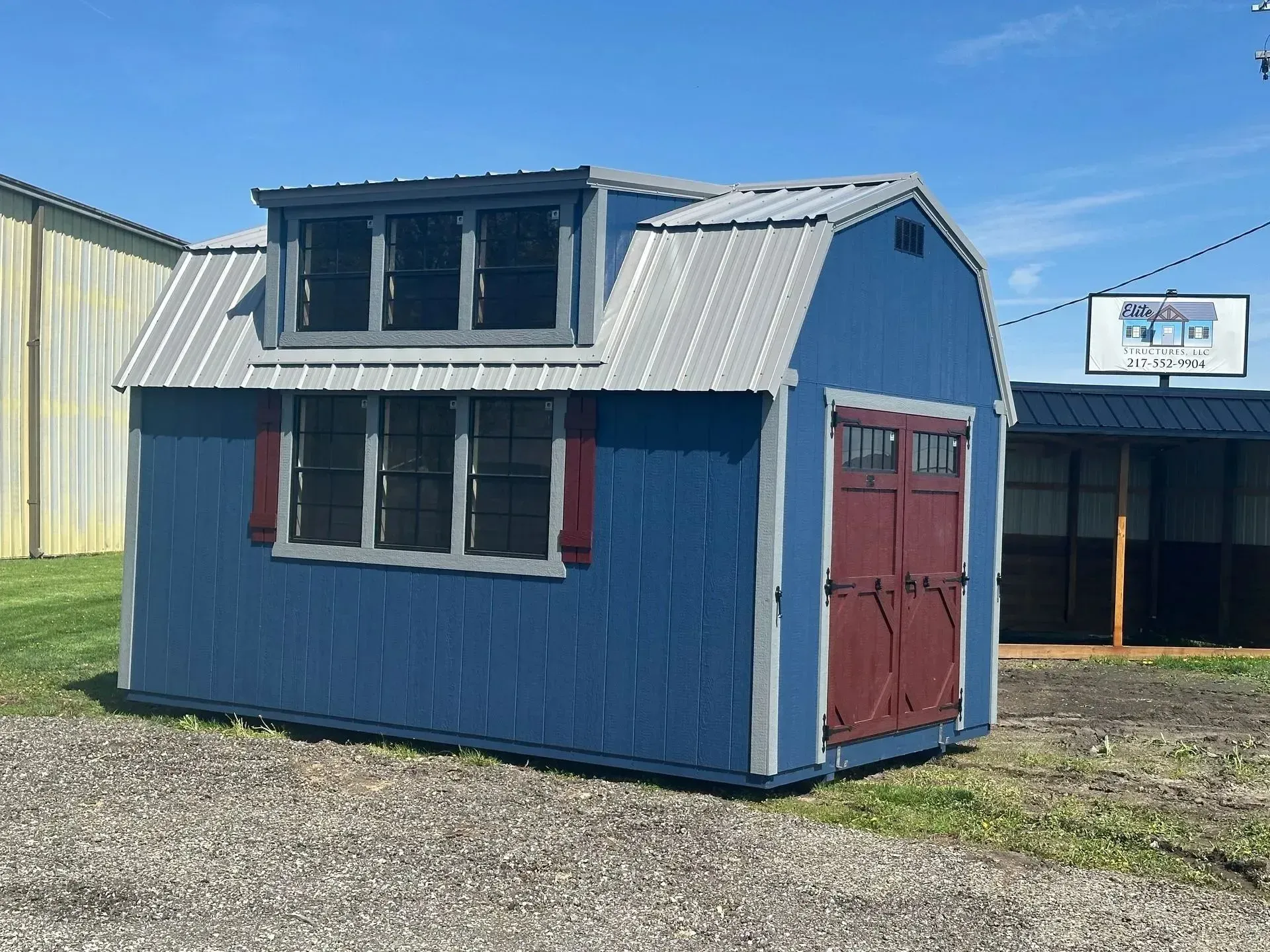 Blue shed with gray metal roof, burgundy door, windows, and trim. Outdoors, sunny day.