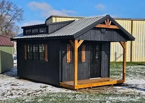 Black cabin with a porch, brown trim, and a small loft under a blue sky.