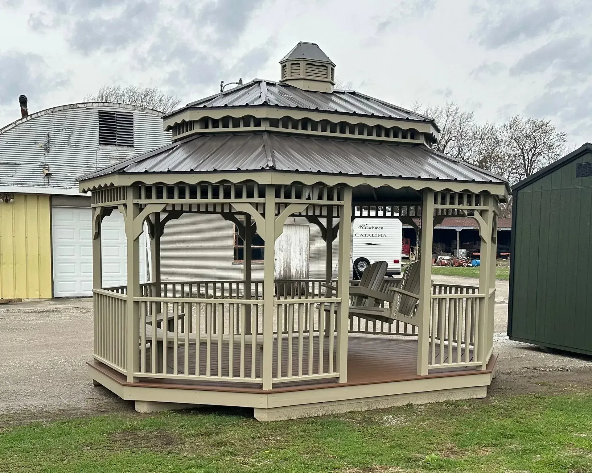 A beige wooden gazebo with a metal roof stands outdoors. In the background are various buildings and greenery.