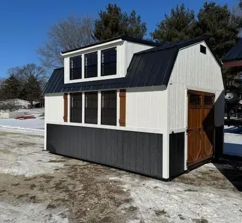 Barn-style storage shed with white and black exterior, brown door, windows, and dark roof on a snowy day.