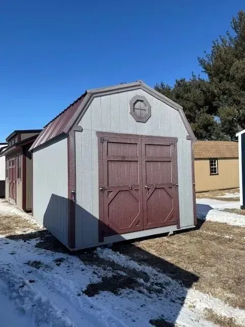 Gray and maroon barn-style shed with double doors, under a blue sky, sitting on snowy ground.