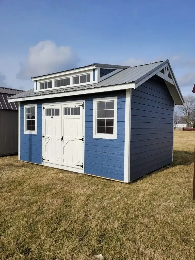 Blue storage shed with white trim, double doors, and dormer windows under a blue sky.