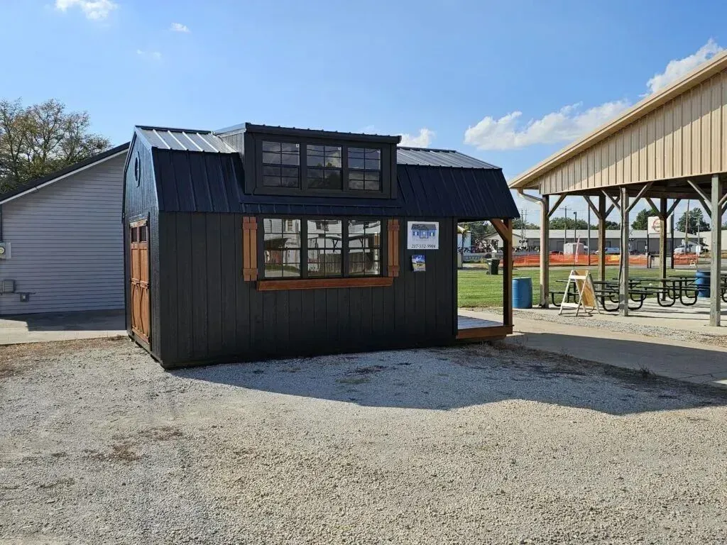 Black shed with wooden trim, small window, and metal roof. Located in a gravel area near a pavilion and building.