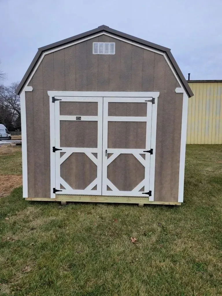Brown barn-style shed with white double doors, white trim, and a small window on a grassy lawn.