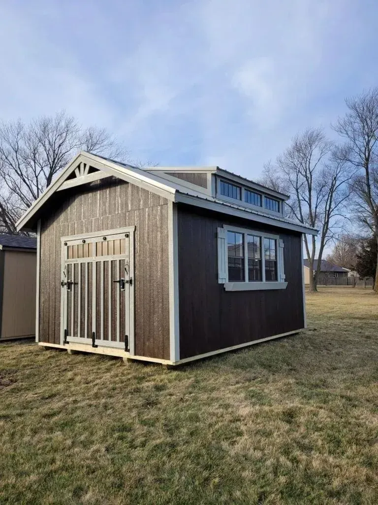 Brown shed with white trim and multiple windows, set in a grassy yard under a blue sky.