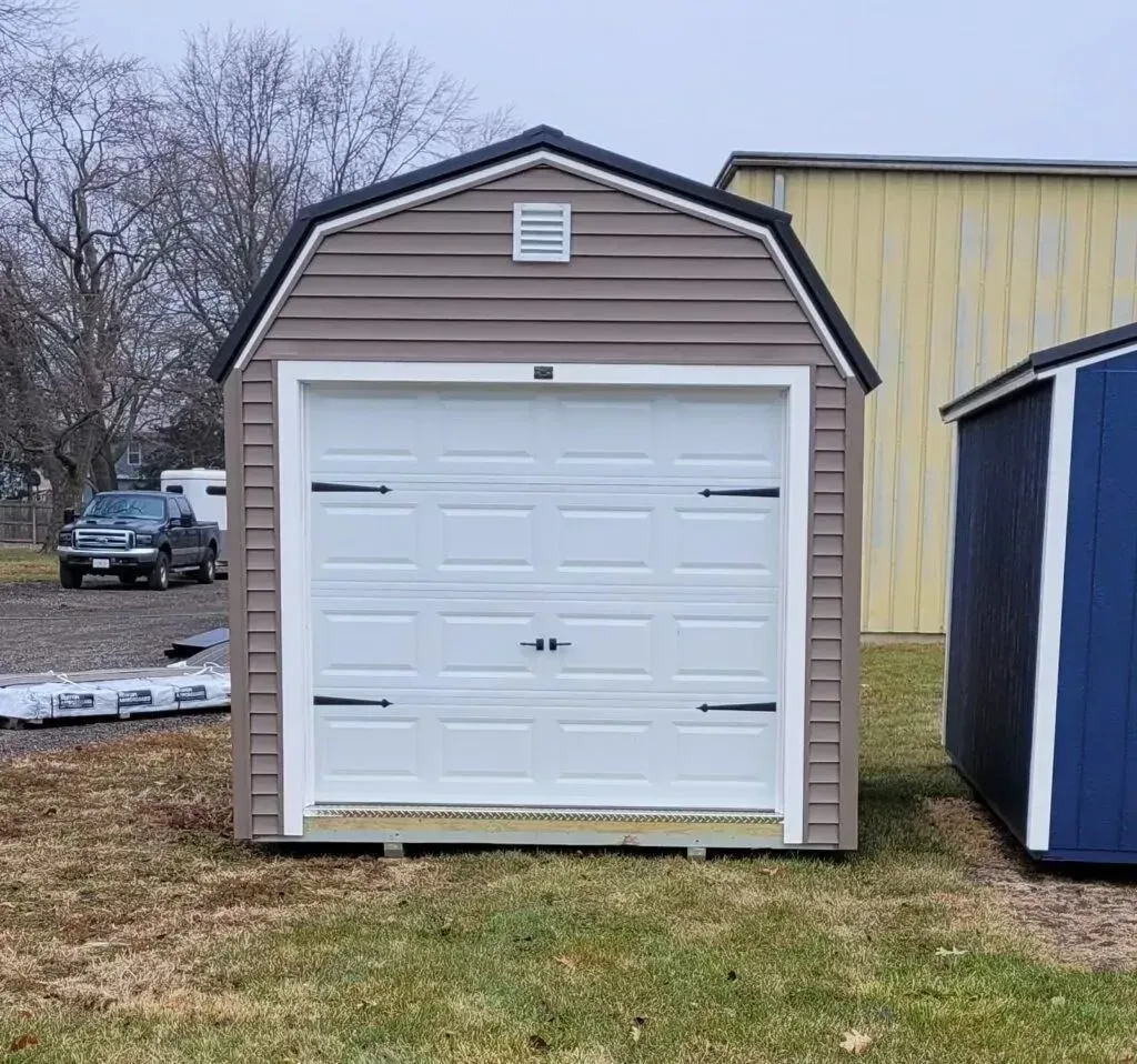 Tan and white shed with a barn-style roof and a garage door, set outdoors on grass.