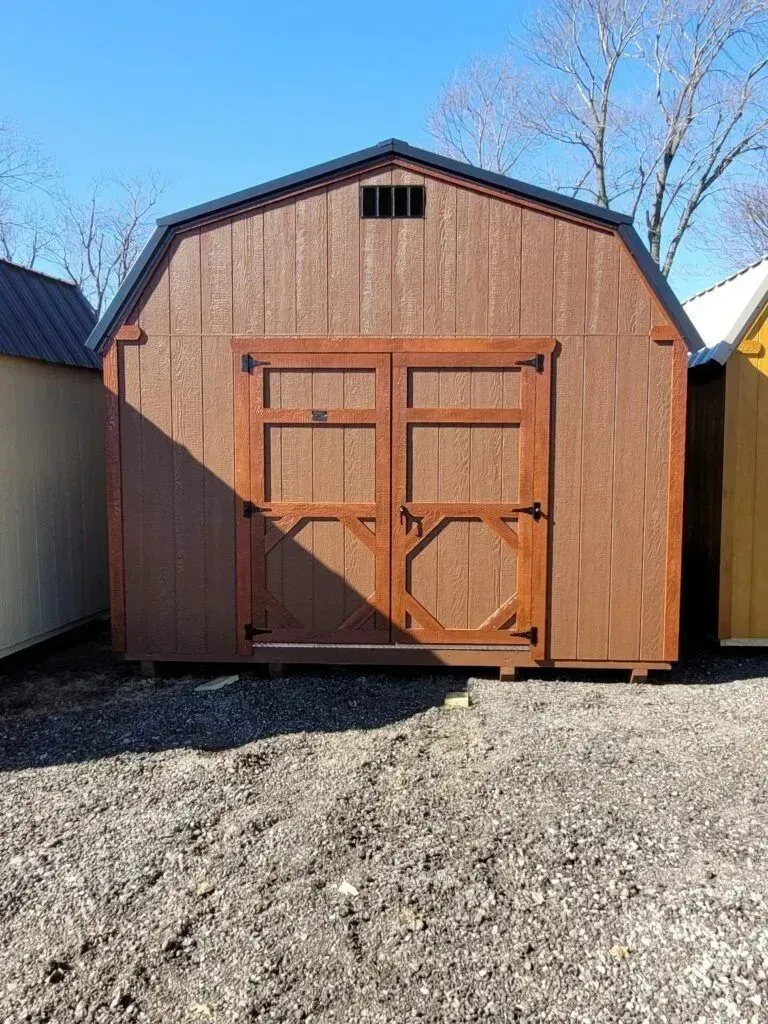 Brown wooden barn-style shed with double doors, vents, and a black roof, set on gravel.