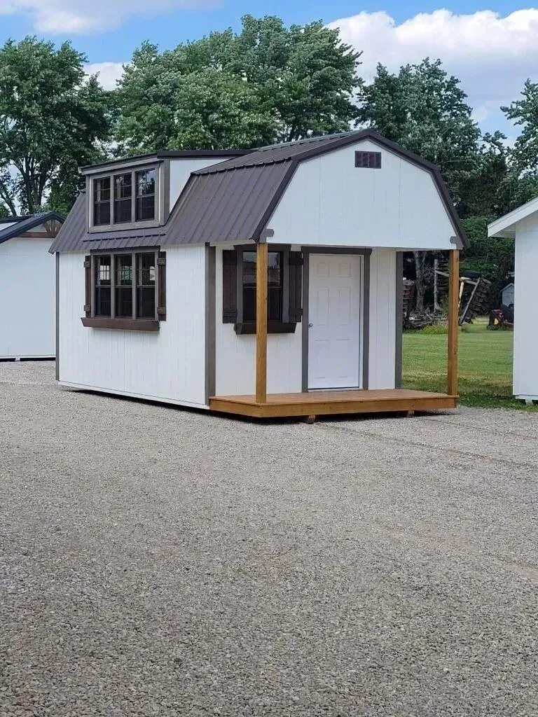 White shed with brown trim and metal roof, with porch. Set on gravel.