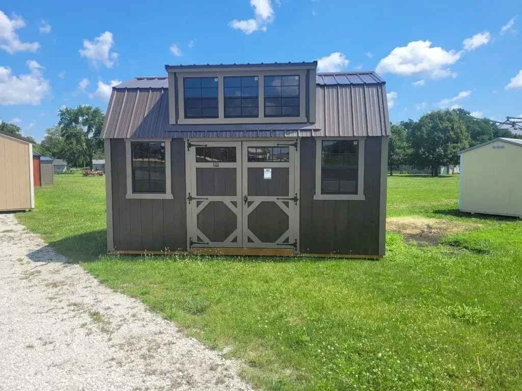 Gray storage shed with dormer, windows, and double doors on a grassy lot.