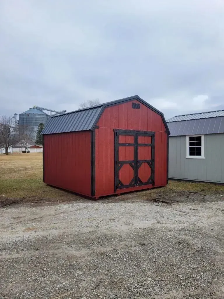 Red barn-style shed with black trim and door, standing on gravel. Another shed visible to the right. Overcast day.