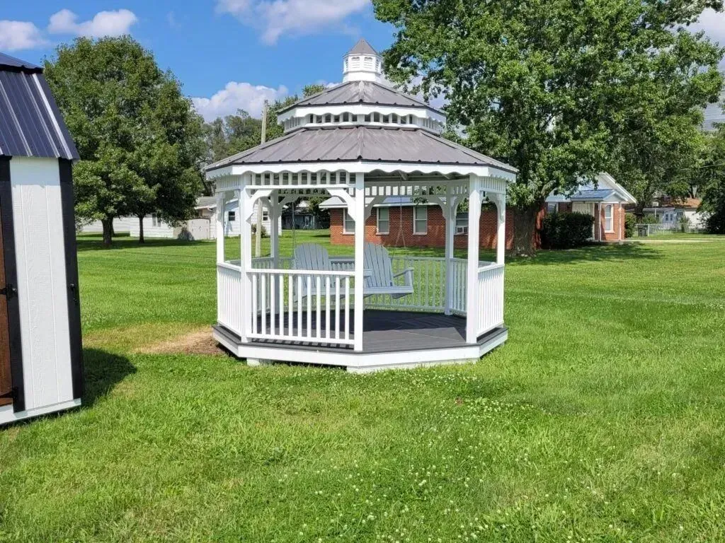 White gazebo with swing seat, in a grassy yard under a blue sky.