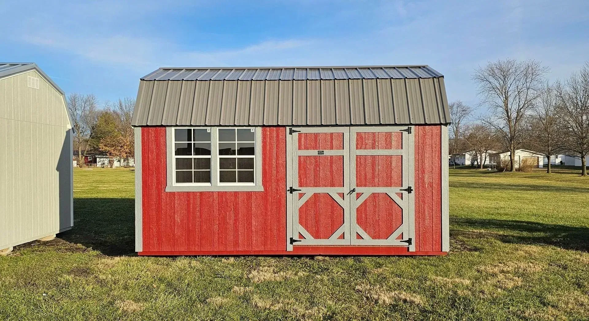 Red barn-style shed with white trim and a gray roof, in a grassy field on a sunny day.