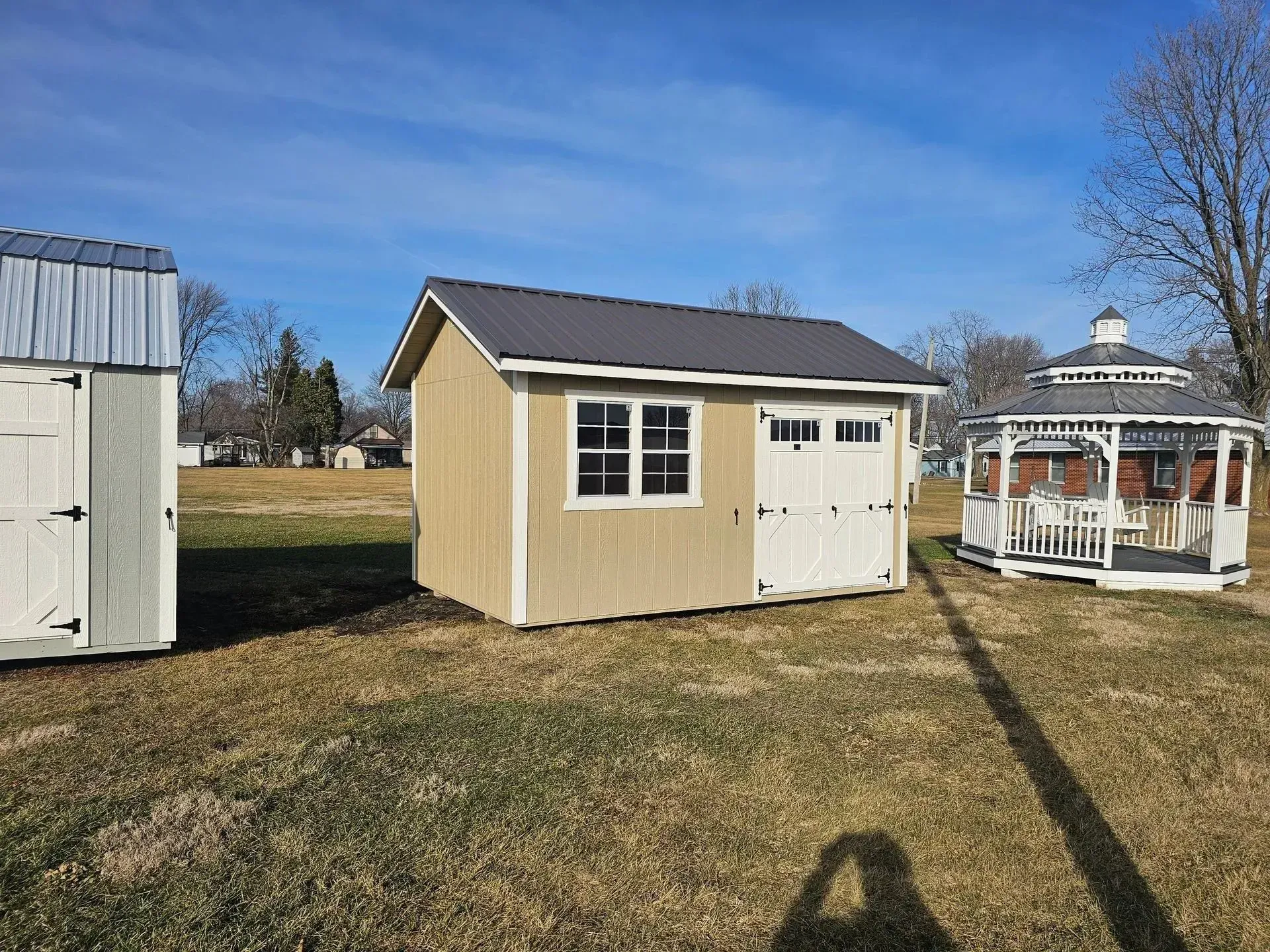 Tan shed with white door and window, grey roof, sitting in a field on a sunny day.