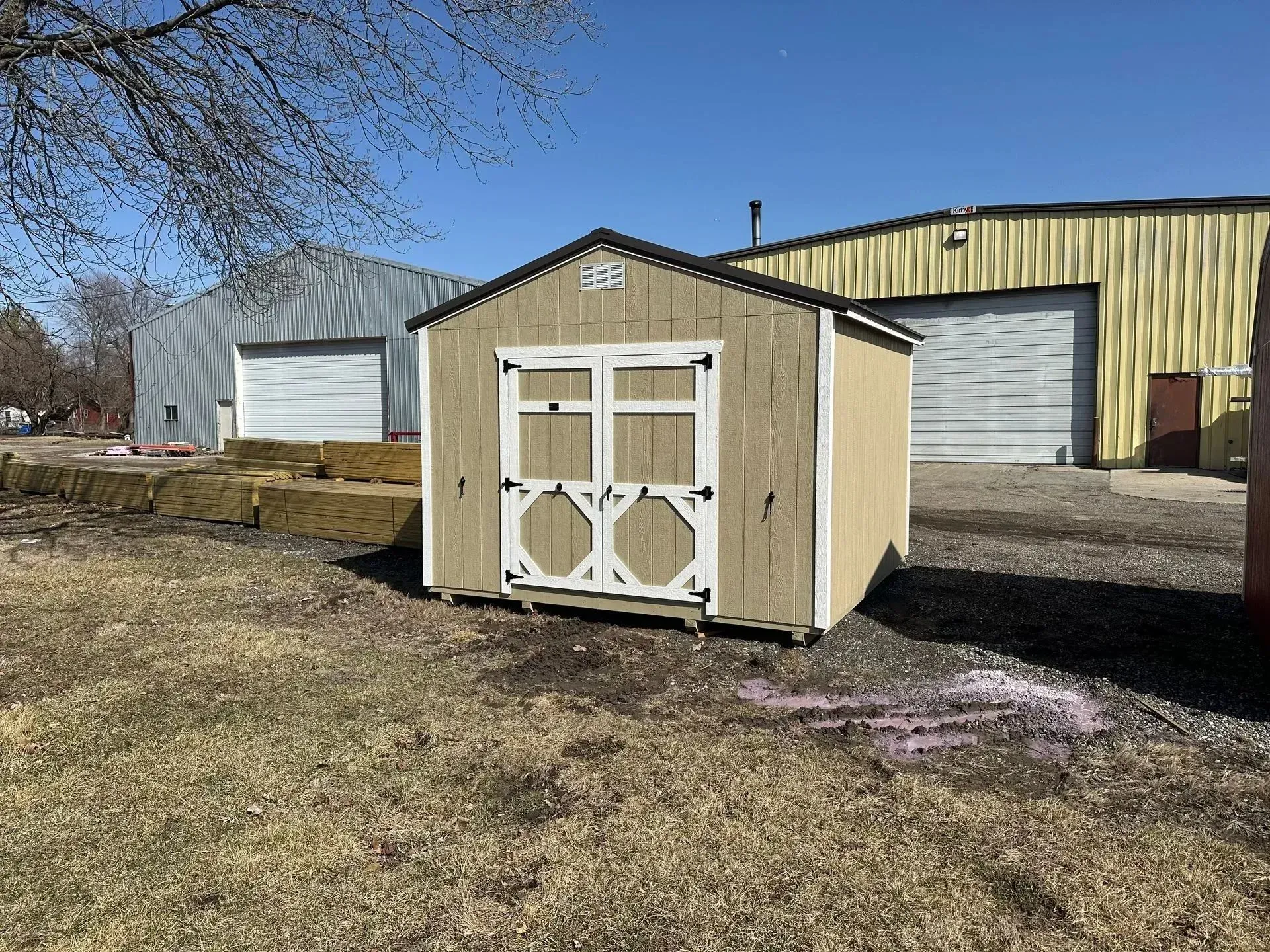 Tan shed with white doors, black trim, and two garage-like buildings in the background on a sunny day.