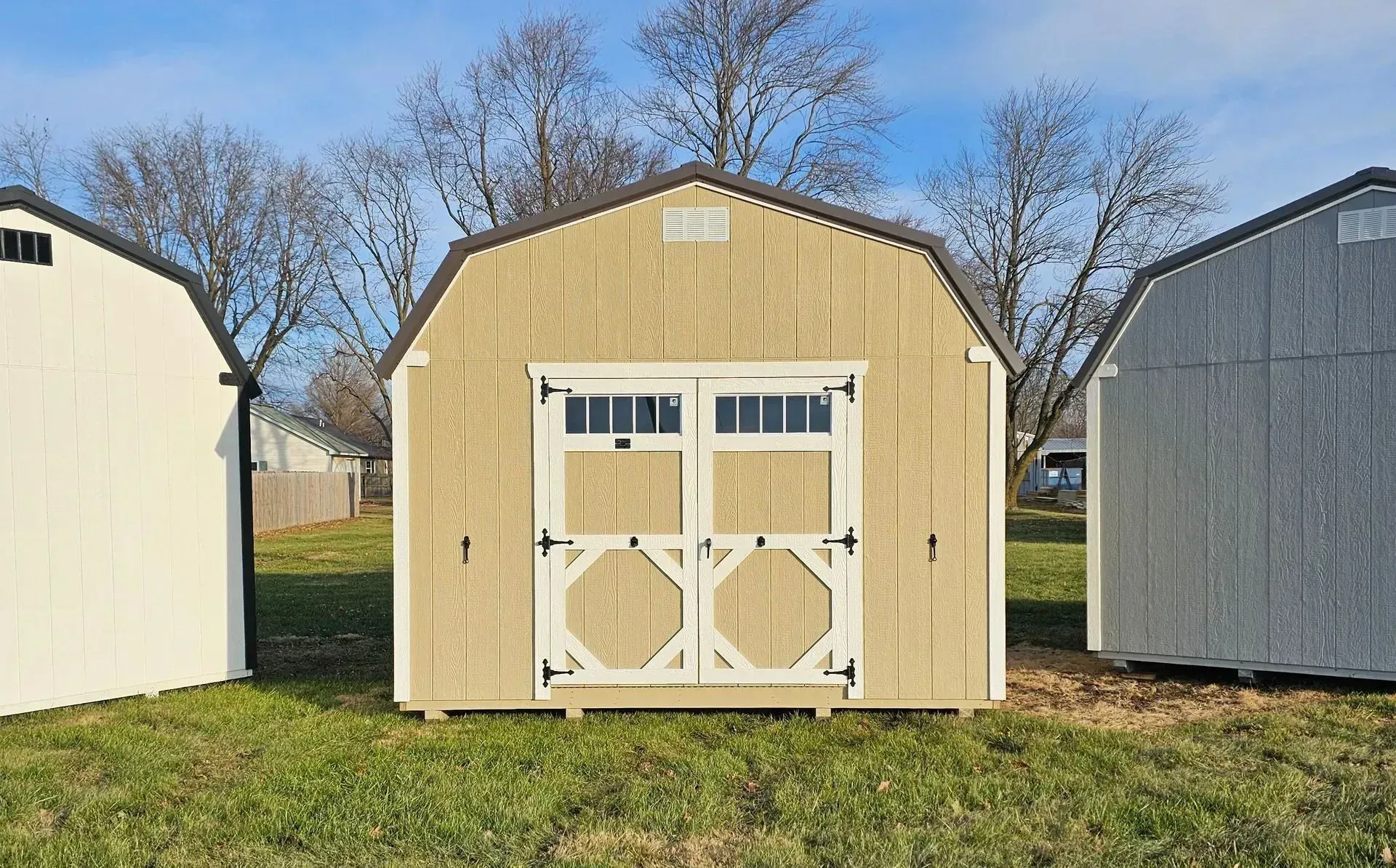 Tan barn-style shed with white trim and double doors on green grass with blue sky background.