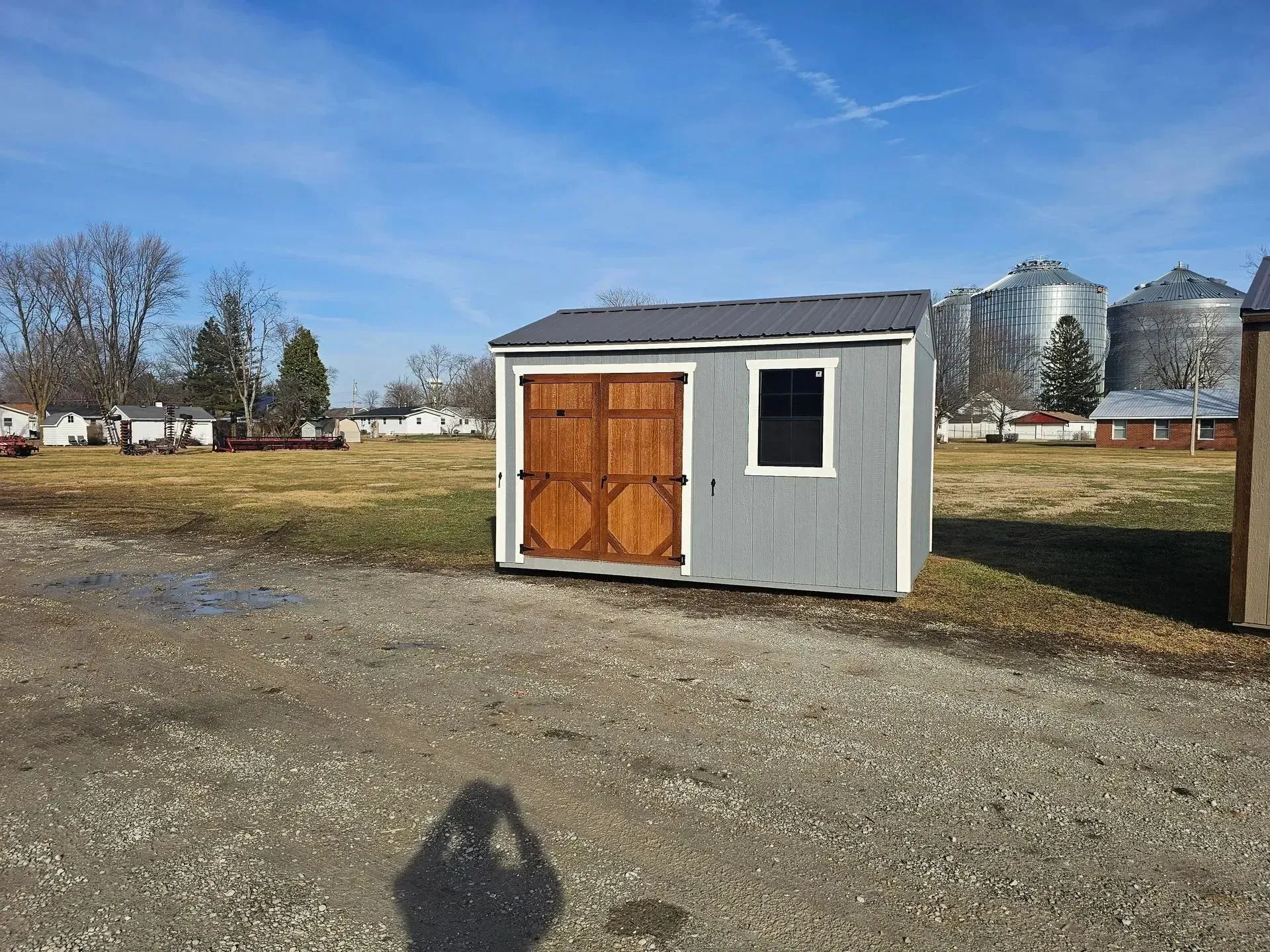 Gray shed with brown doors and small window, gravel ground, field, blue sky, silos in background.