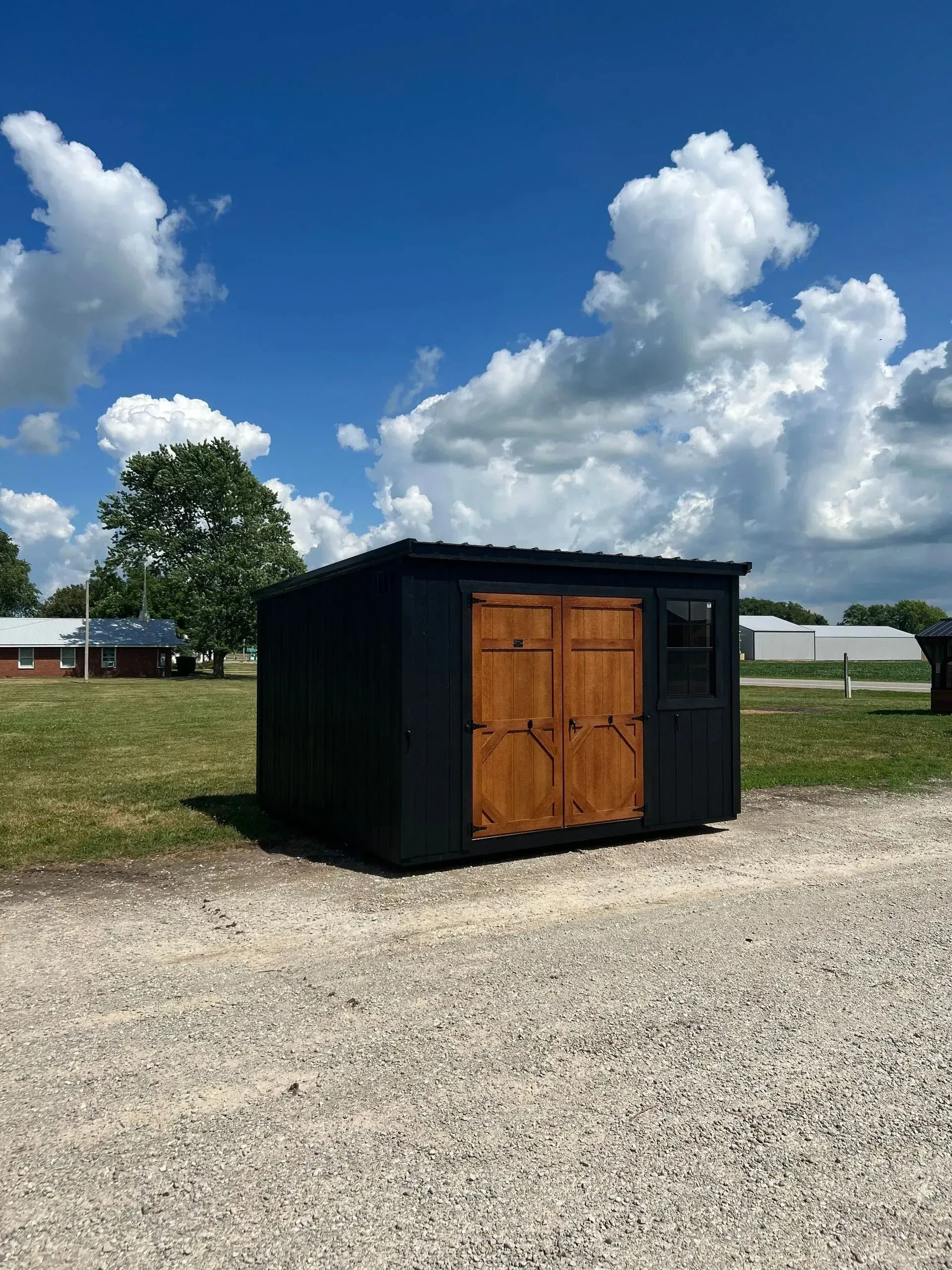 Black shed with brown wooden double doors, set on a gravel path under a blue sky with clouds.
