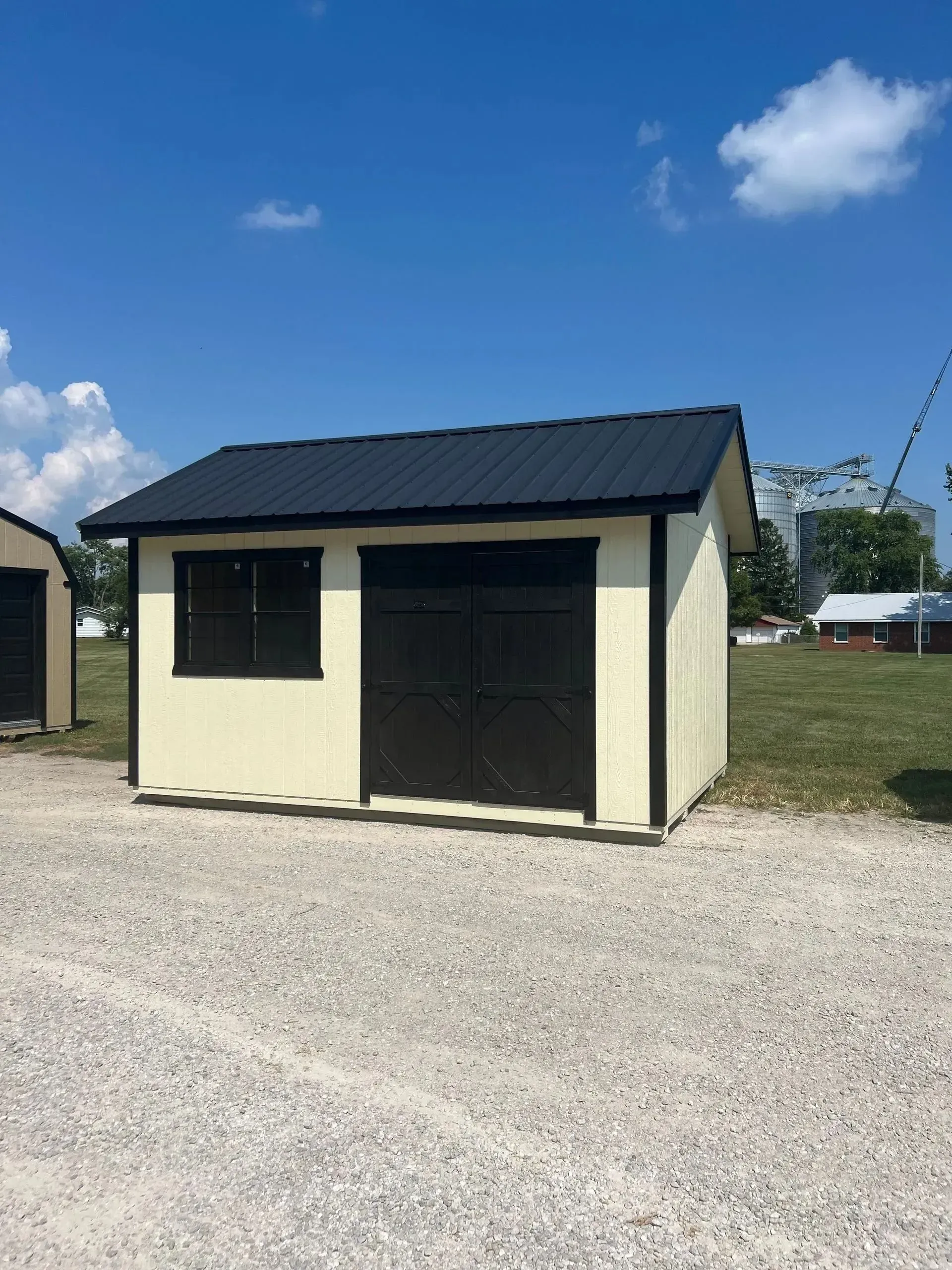 Cream-colored shed with black door, window, and roof, set on gravel with a clear blue sky.